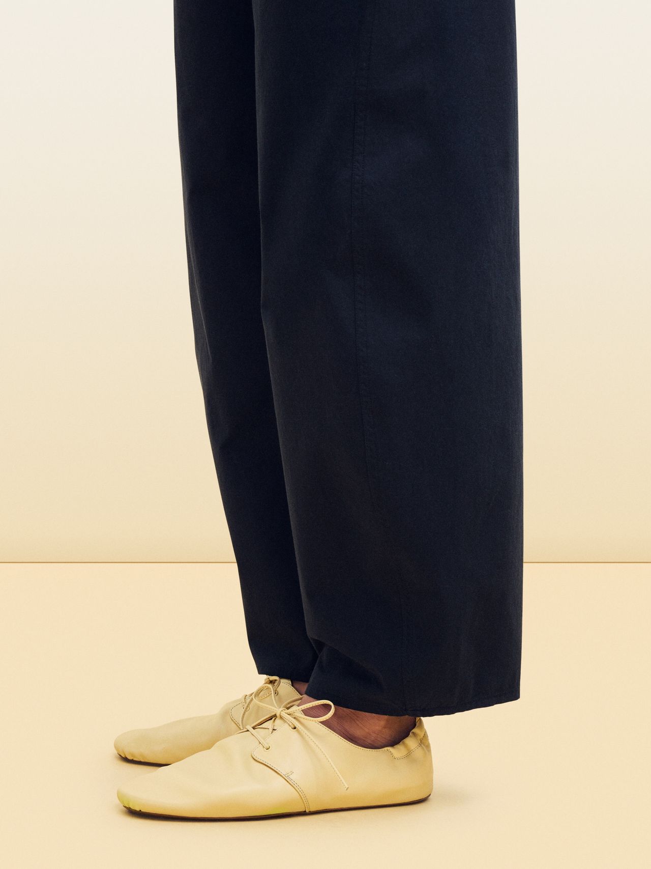 Person wearing loose black pants and beige flat shoes with laces, standing on a light beige surface, against a gradient background.