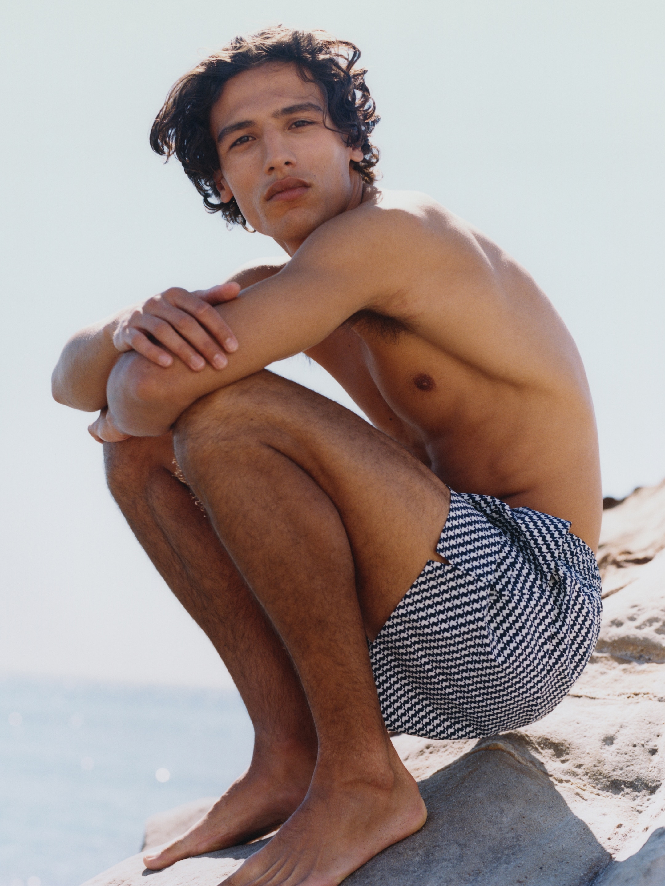 A person with curly hair sits on a rock by the sea, wearing patterned swim shorts, under a clear blue sky.