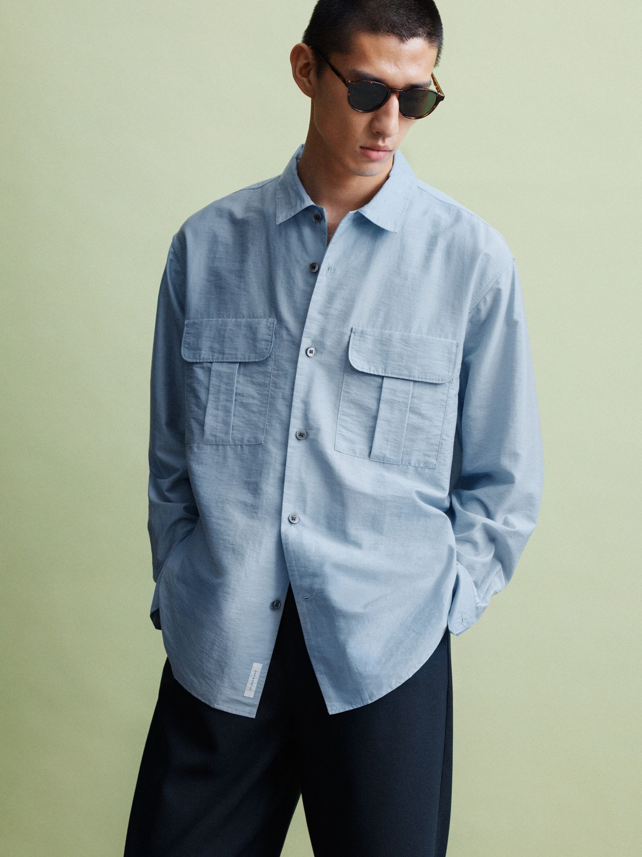 Man wearing sunglasses, a light blue button-up shirt with pockets, and dark trousers, standing against a light green background.