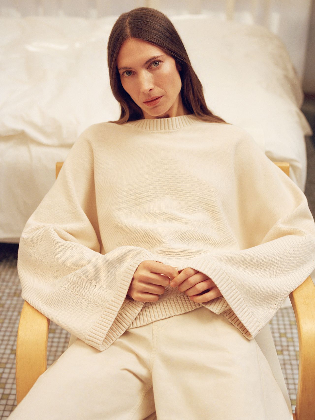 Person with long brown hair in a cream sweater and pants sits on a chair in a softly lit room with a tiled floor and bed in the background.