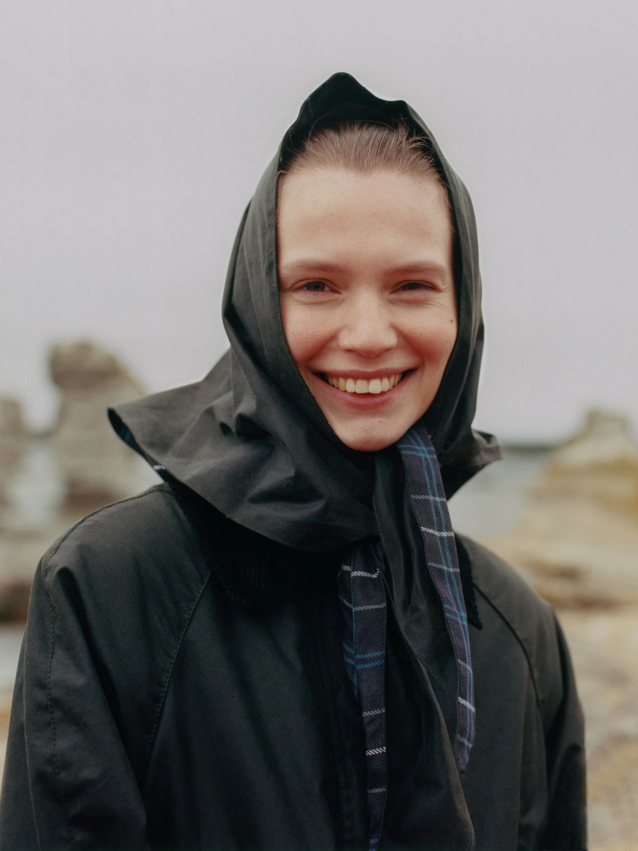A person smiling, wearing a dark headscarf and coat, stands outdoors with blurred rocky formations in the background.