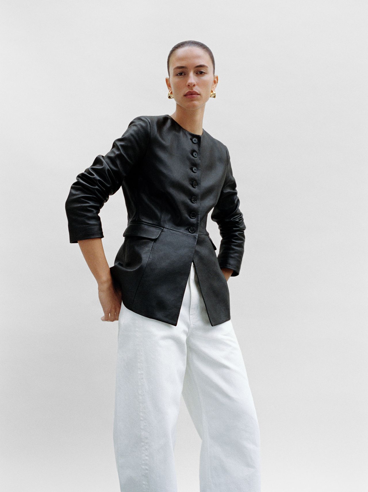 Person wearing a black leather jacket and white pants, standing against a plain background, looking confidently at the camera.