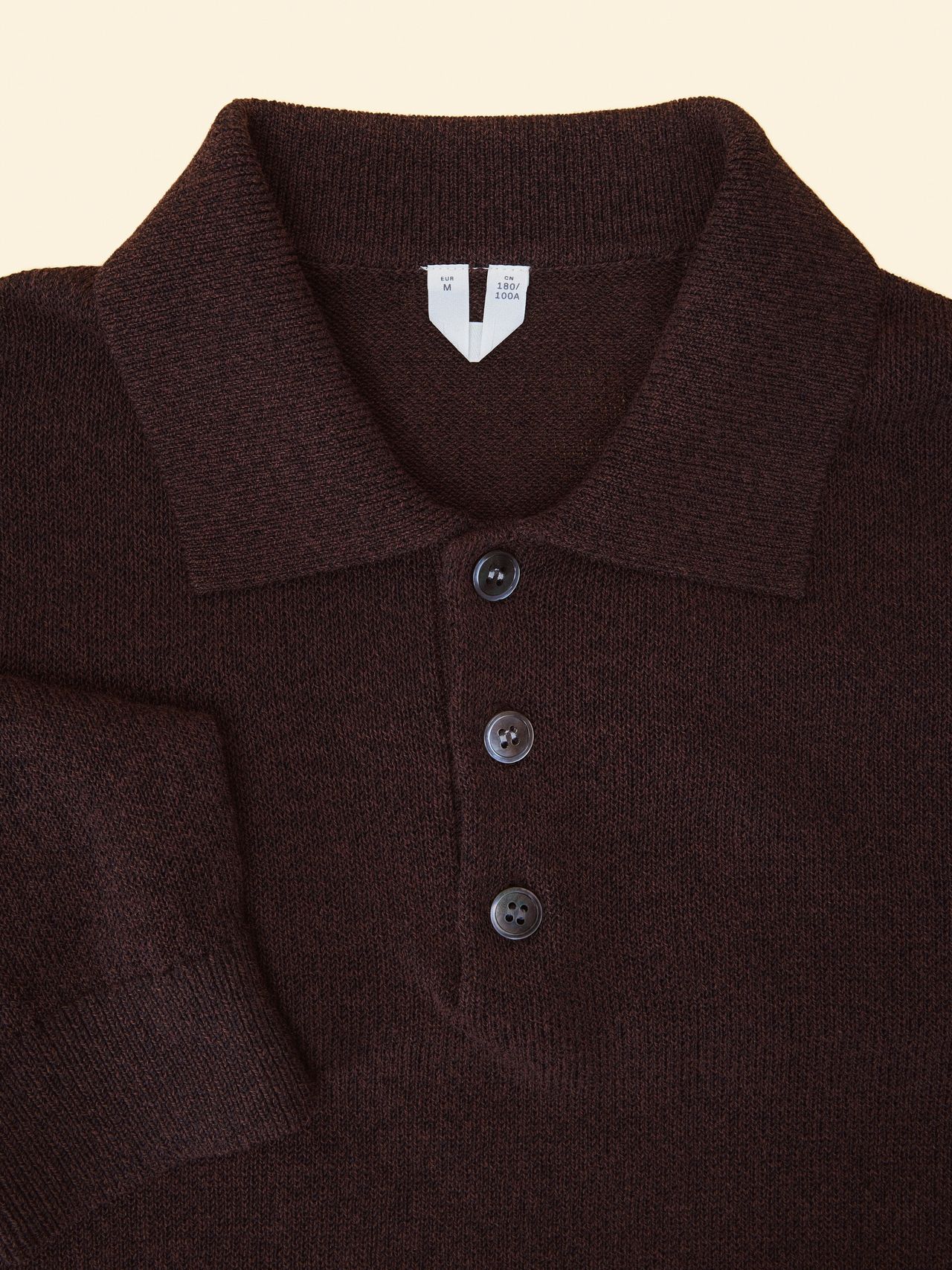 Dark brown, long-sleeve polo shirt with three buttons and a folded collar on a beige background, displaying a soft, textured fabric.