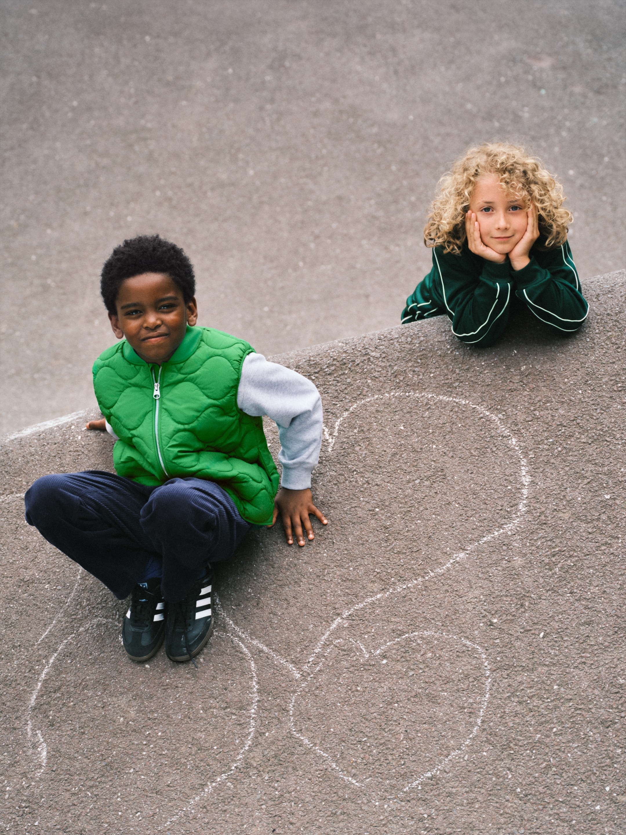 Two children on concrete; one sits on a chalk heart drawing, wearing a green vest, the other leans on a wall, hands on cheeks, with curly hair.