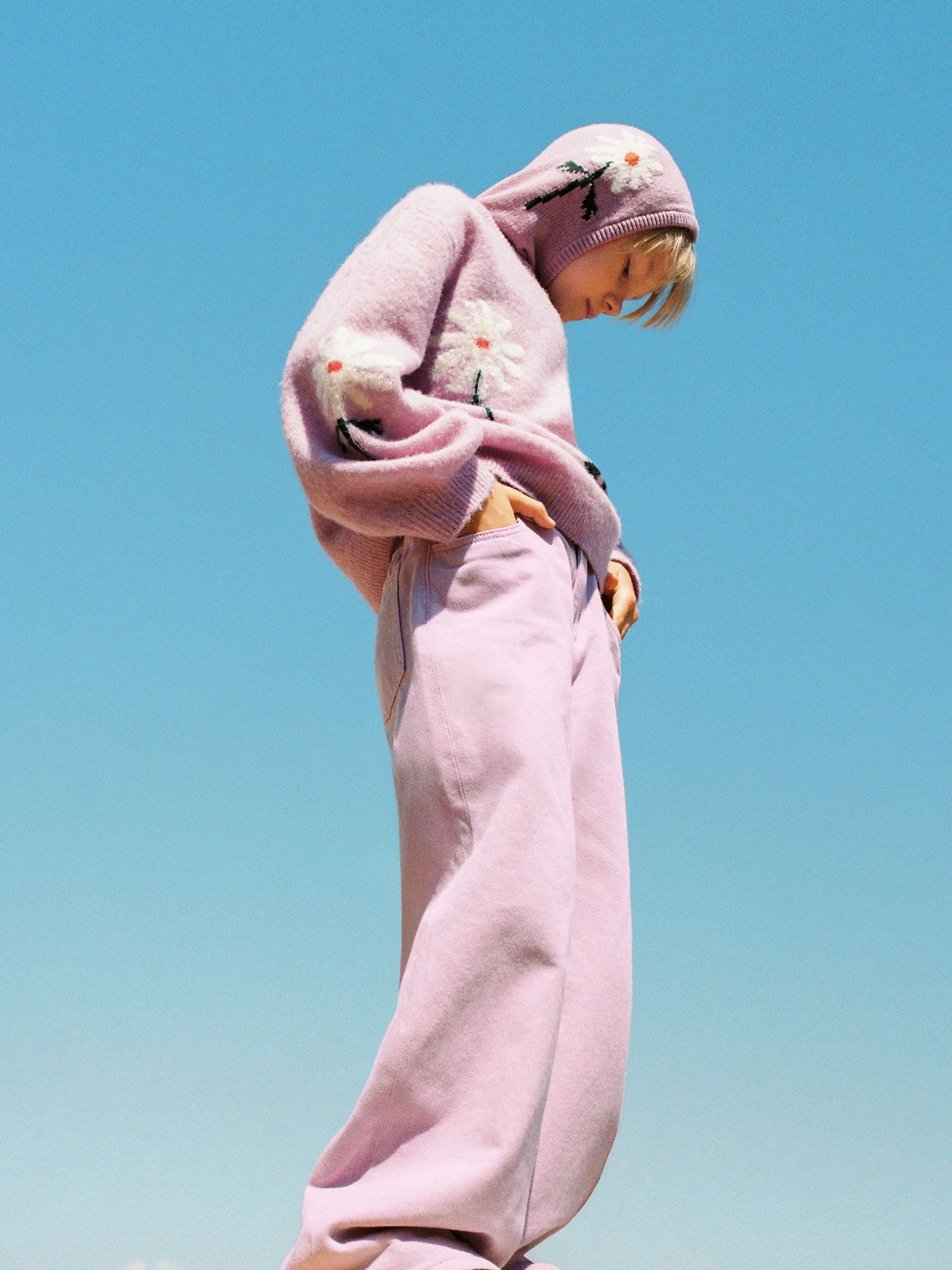 Person in a pastel pink outfit with floral embroidery stands against a clear blue sky, looking down with hands in pockets.