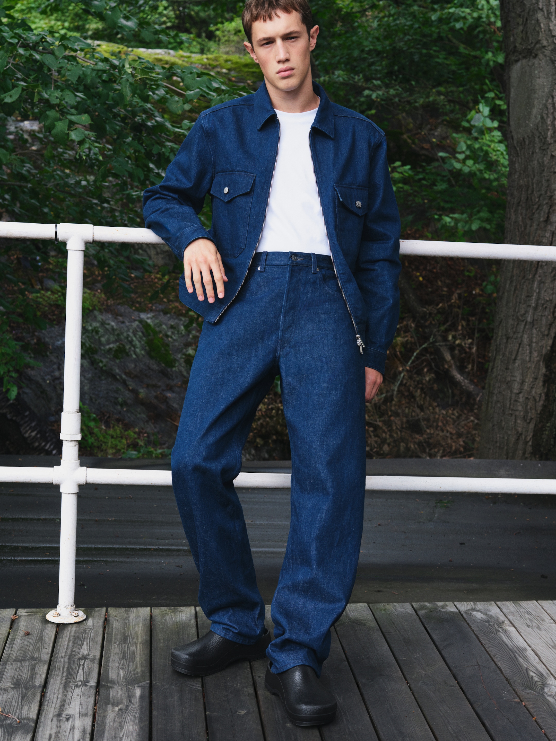 Person in a denim jacket and jeans, white shirt, and black shoes, standing on a wooden deck with greenery in the background.