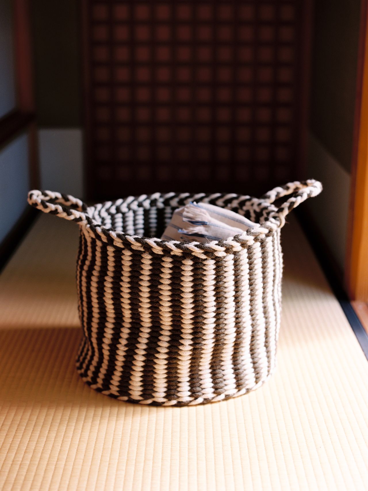 A black and white striped woven basket with handles sits on a tatami mat, containing folded items, in a softly lit room.