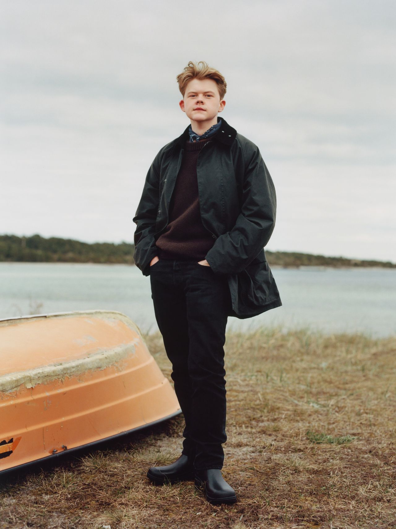 Young person in a dark coat and boots stands confidently by an overturned boat on a grassy shore, with a cloudy sky and water in the background.
