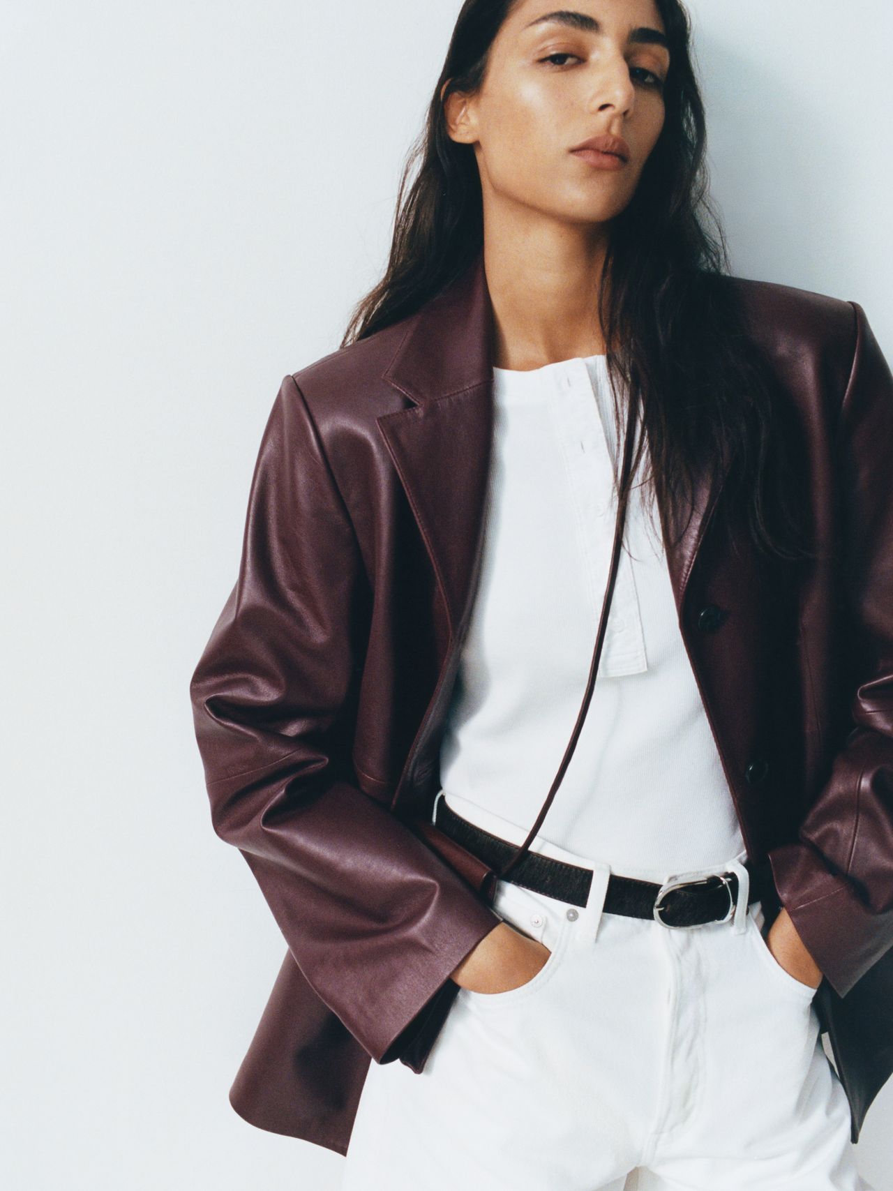 Person wearing a burgundy leather jacket over a white shirt and pants, hands in pockets, standing against a light background.
