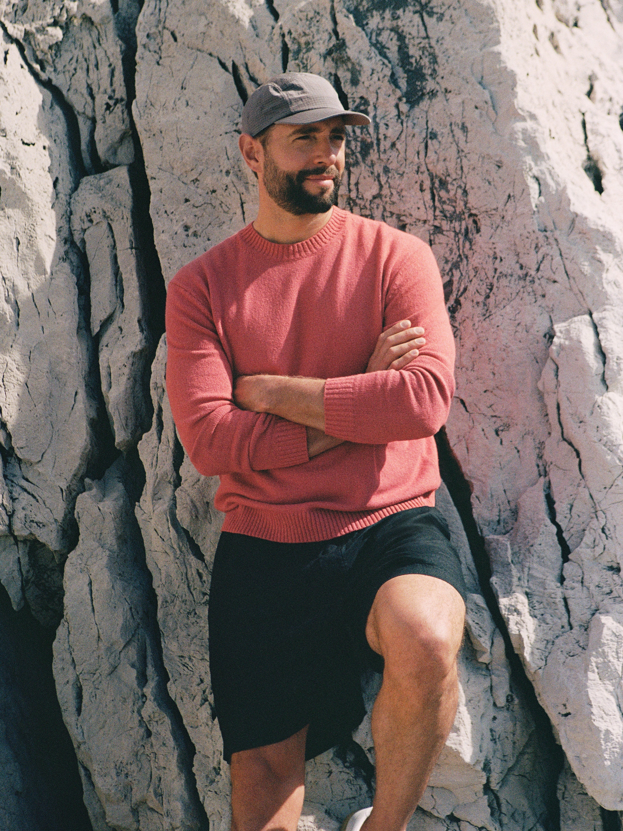 Man in a pink sweater and cap, sitting against a large rock, arms crossed, looking into the distance.