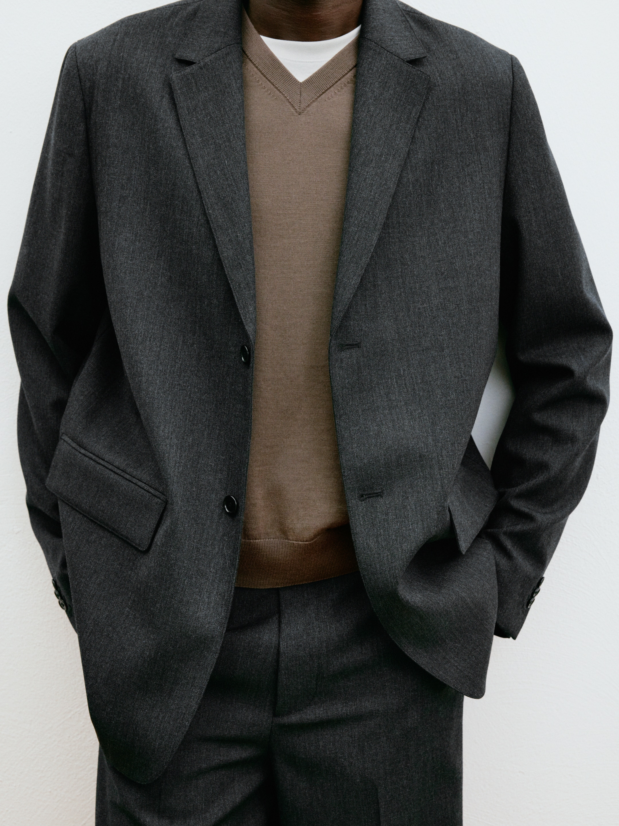 Person wearing a dark gray suit over a brown V-neck sweater and white shirt, with hands in pockets against a plain background.