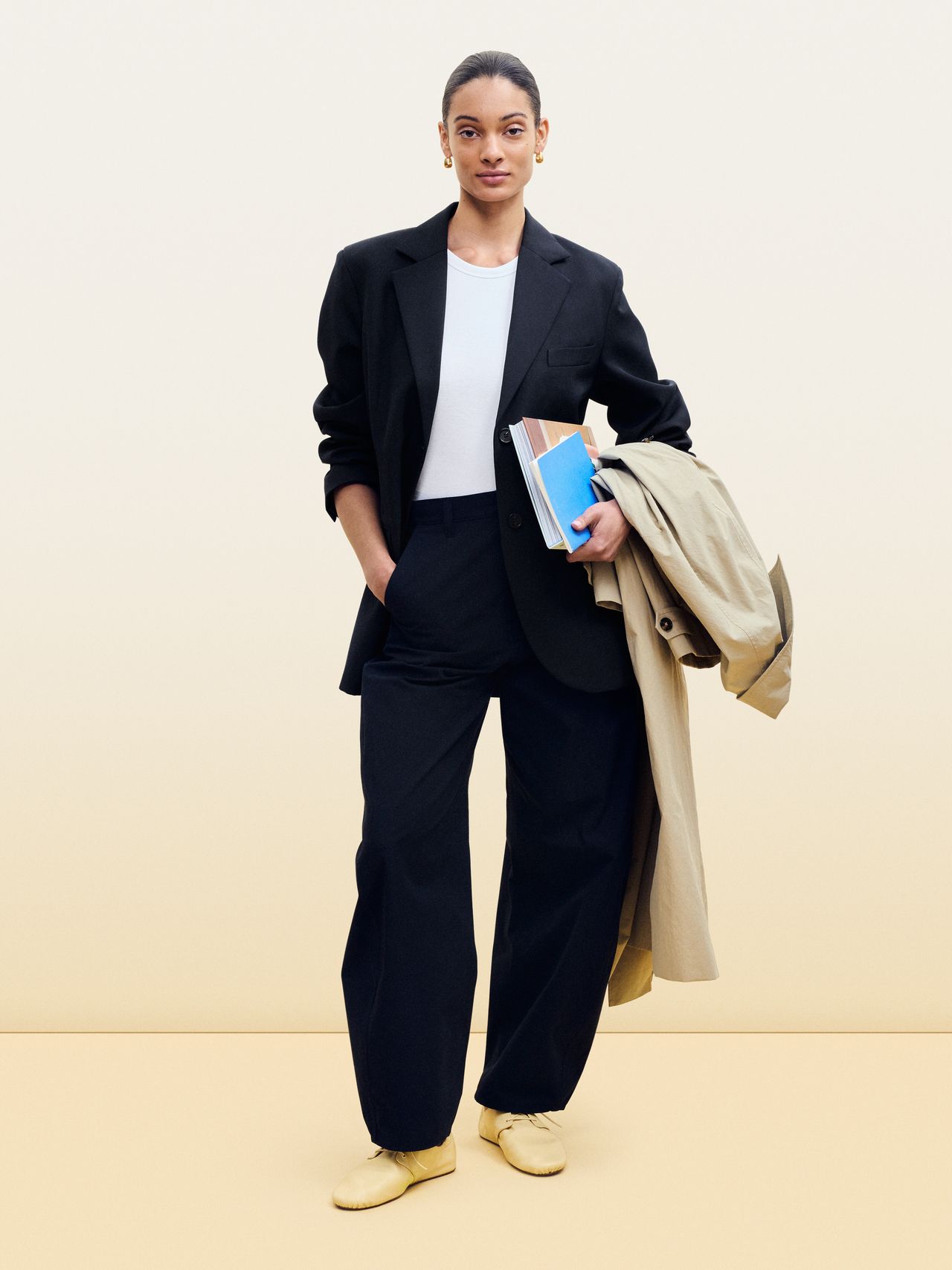 Person in a black suit and white shirt holding books and a beige coat, standing against a neutral background.