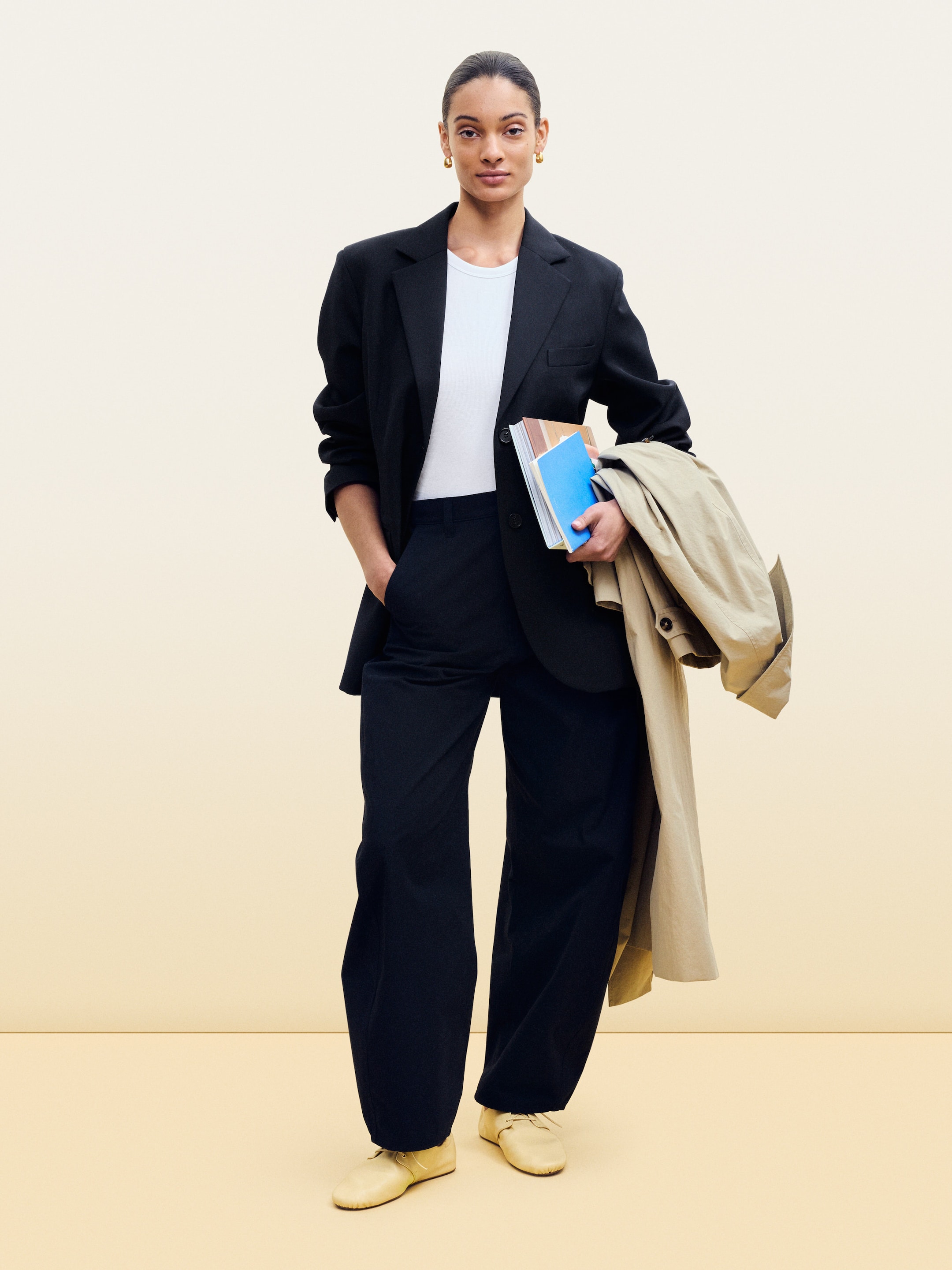 Person in a black suit and white shirt holding books and a beige coat, standing against a neutral background.