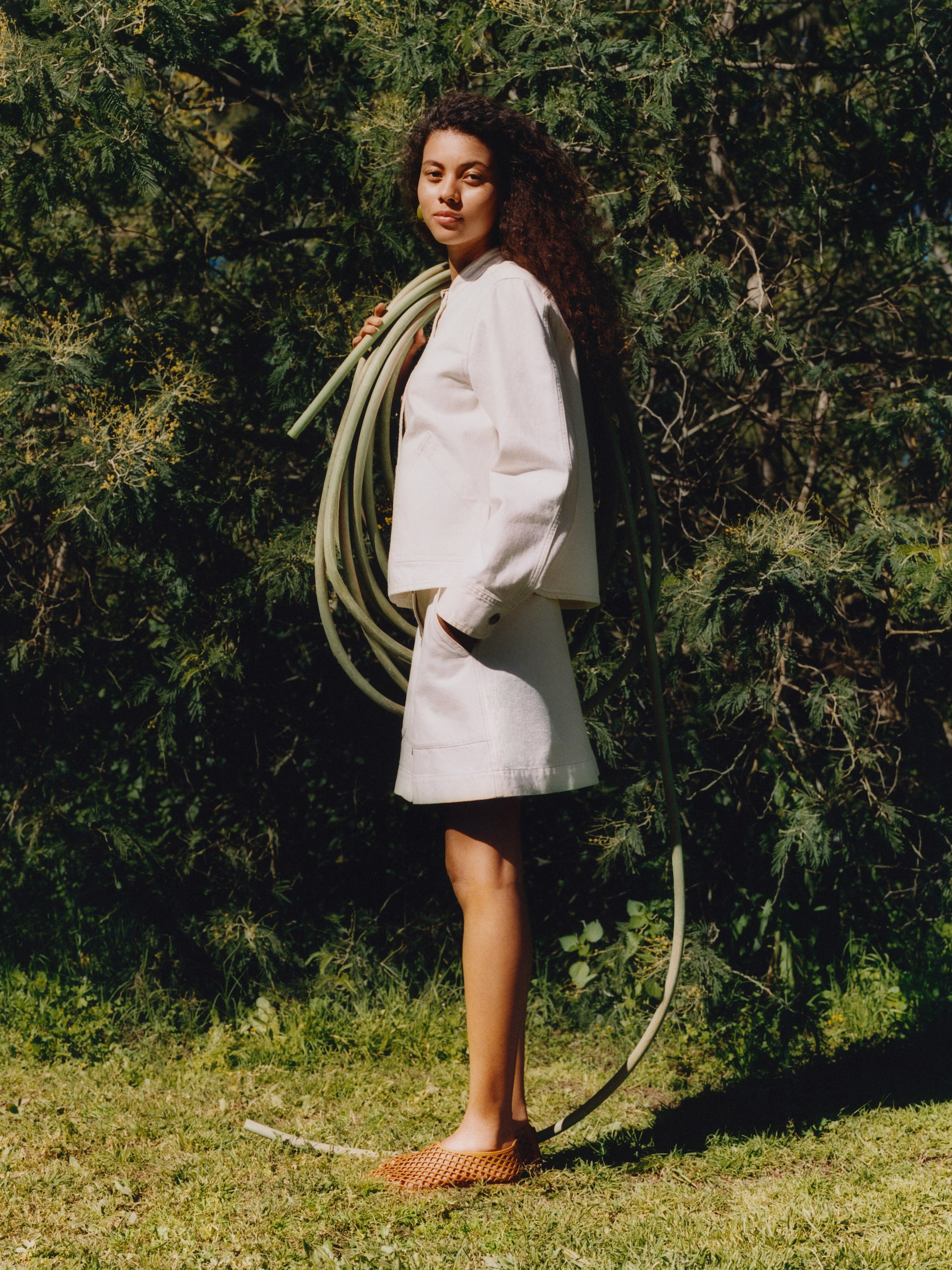 Woman with curly hair stands on grass, wearing a light outfit, holding a coiled garden hose over her shoulder, with trees in the background.