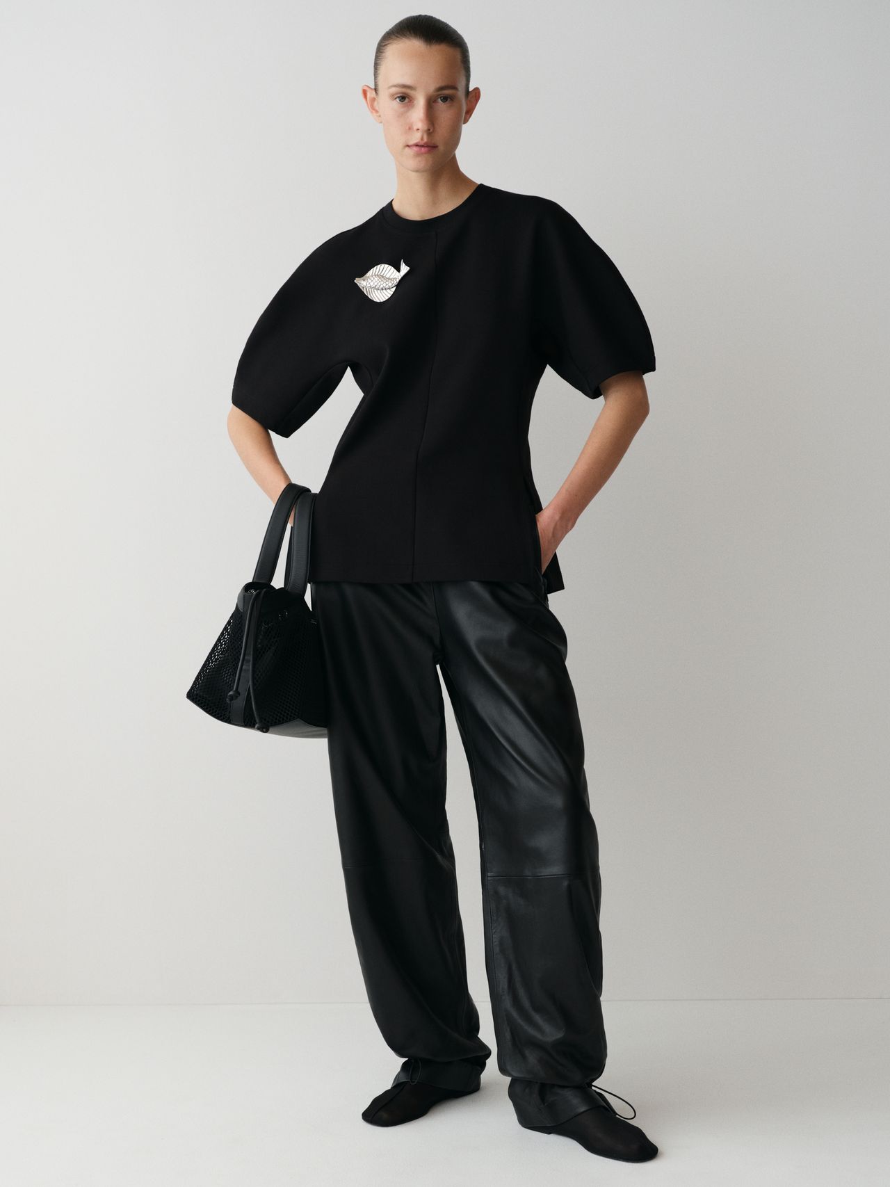 Person in black oversized top and leather pants, holding a textured black bag, standing against a plain white background.