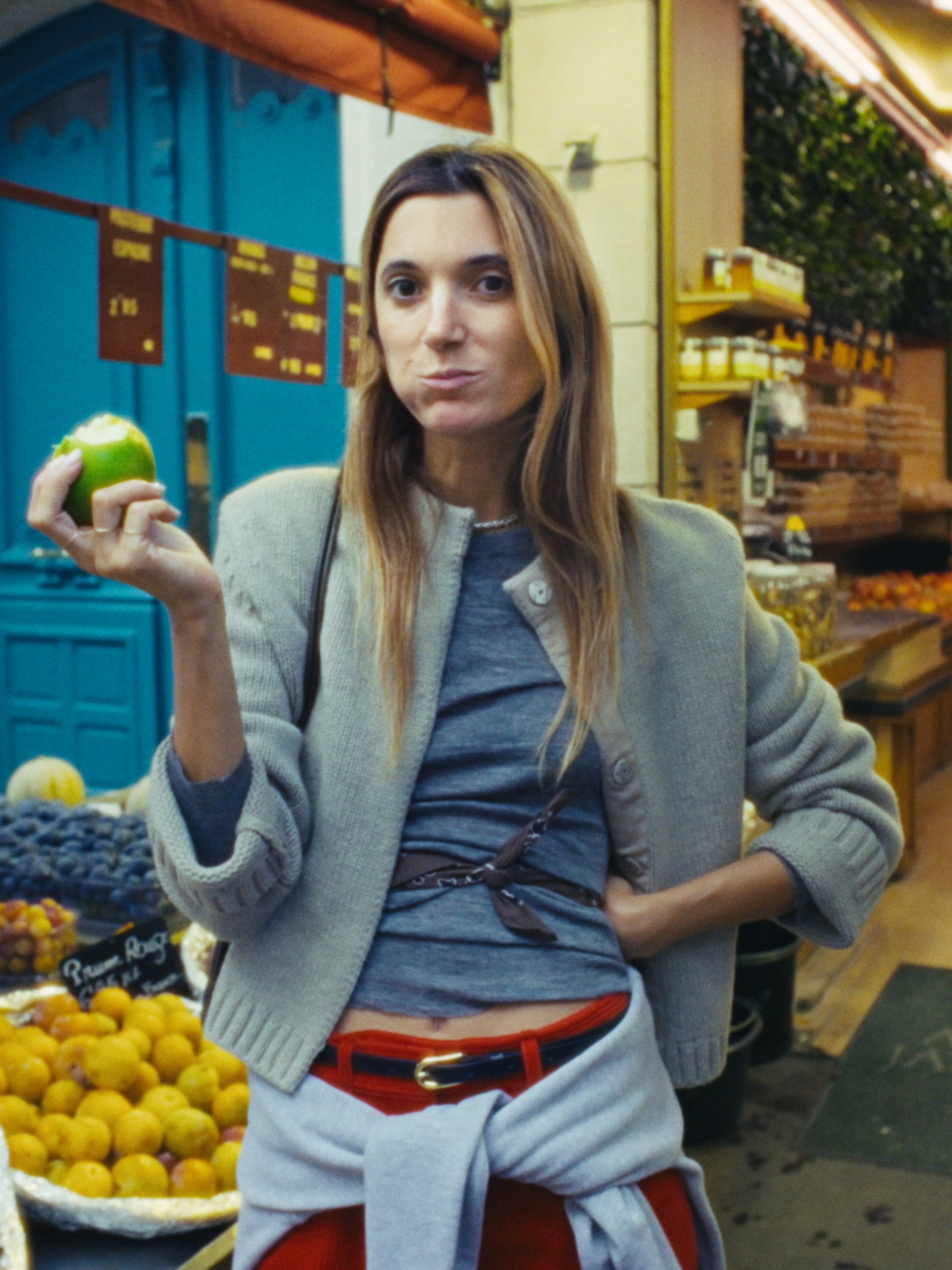 A woman in a market, wearing a gray cardigan and red pants, holds a green apple with a bite taken out of it, standing near a fruit display.