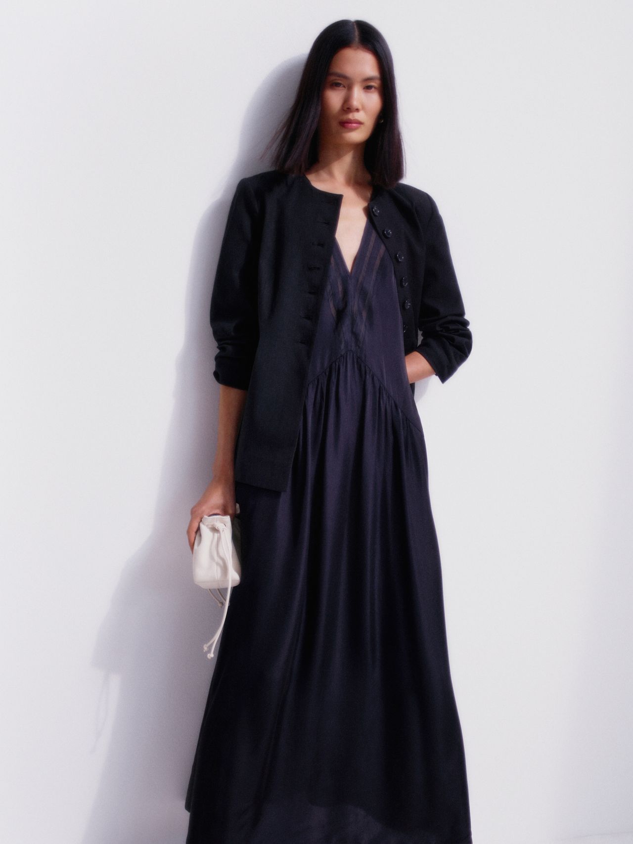 Woman in a black dress and jacket holding a small white purse, standing against a plain white background.