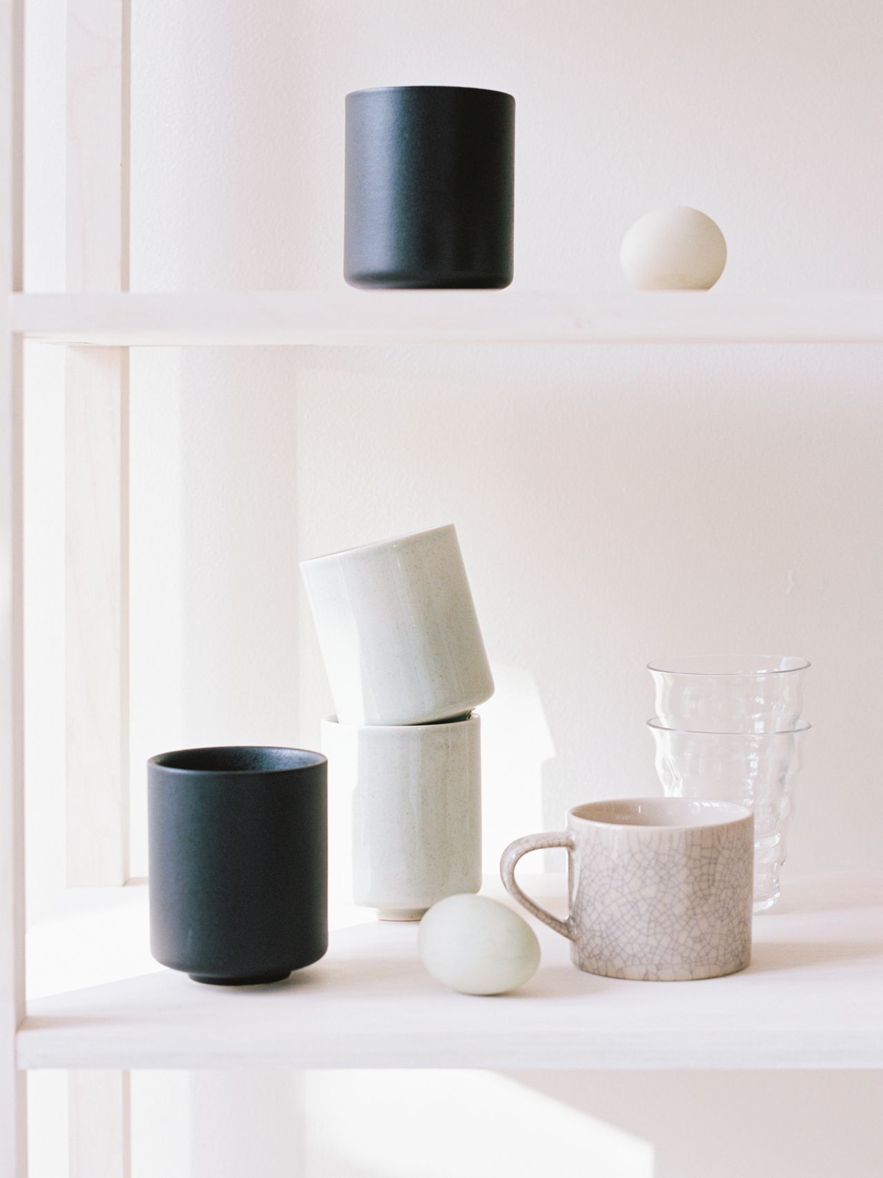 Minimalist shelf display with stacked cups, a mug, small round objects, and two clear glasses, all arranged on light wooden shelves.