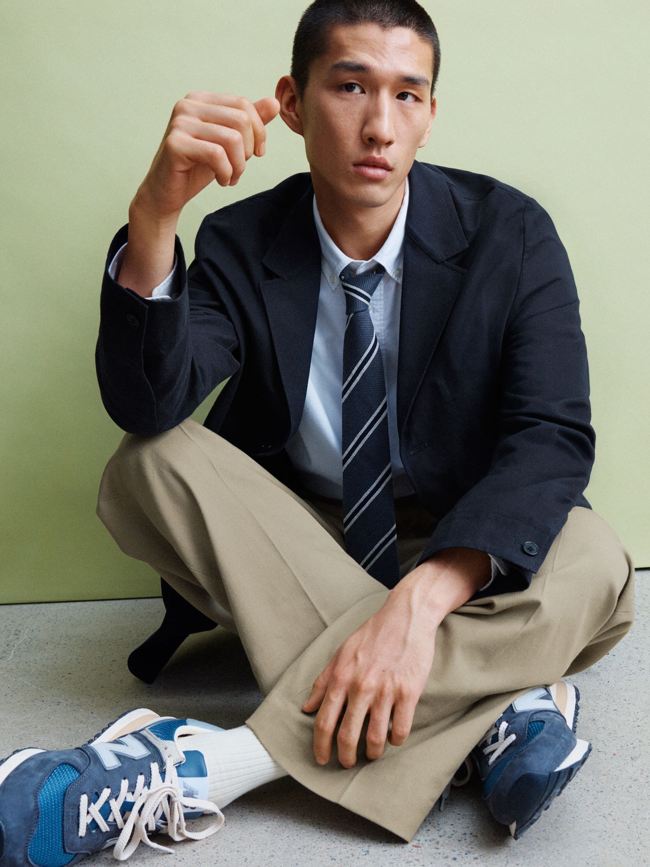 Person seated on the floor, wearing a black blazer, striped tie, khaki pants, and blue sneakers against a light green background.
