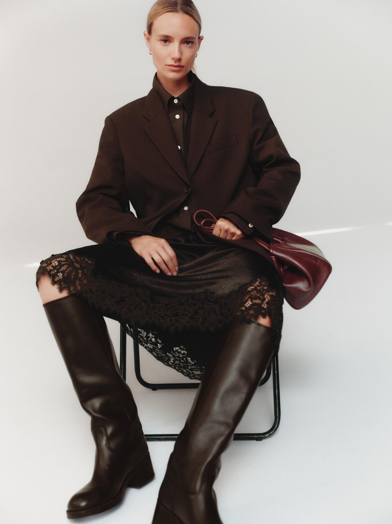 A person sits on a chair wearing an oversized brown blazer, black lace skirt, and knee-high boots, holding a maroon handbag.