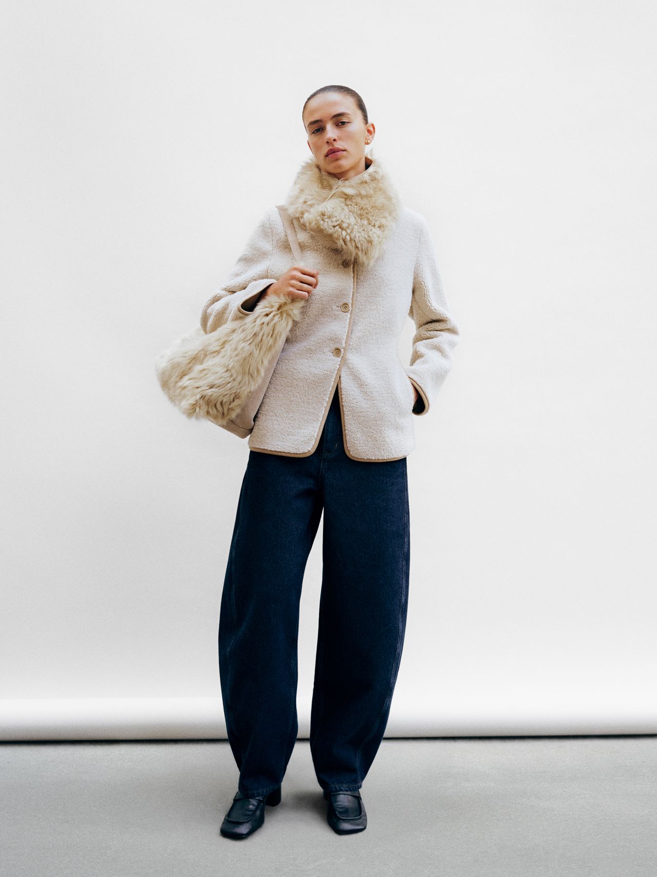 Person in a beige textured coat with fur collar, holding a matching bag, wearing wide-leg dark pants and black shoes, standing against a plain backdrop.