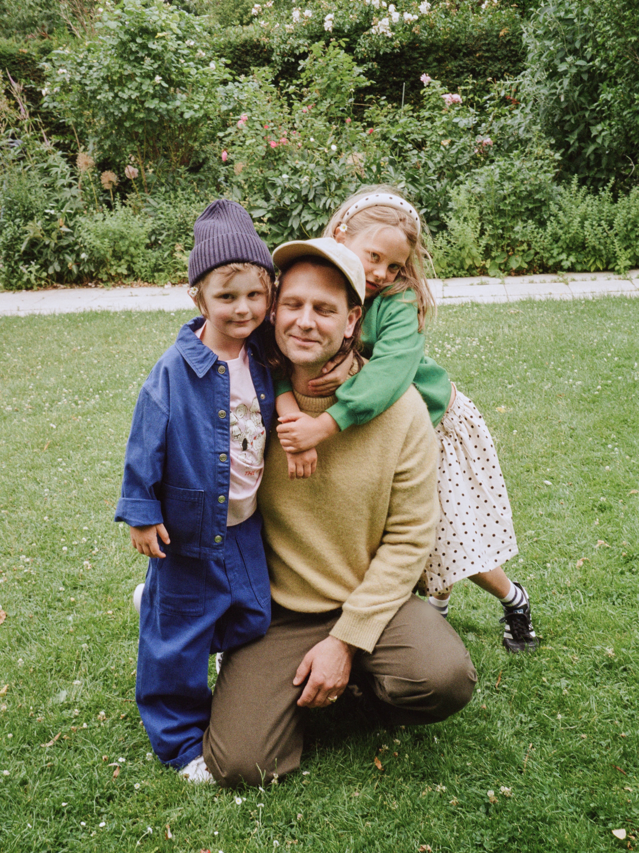 A man kneels on the grass, smiling with his eyes closed, hugged by two children, one on each side, surrounded by greenery and flowers.