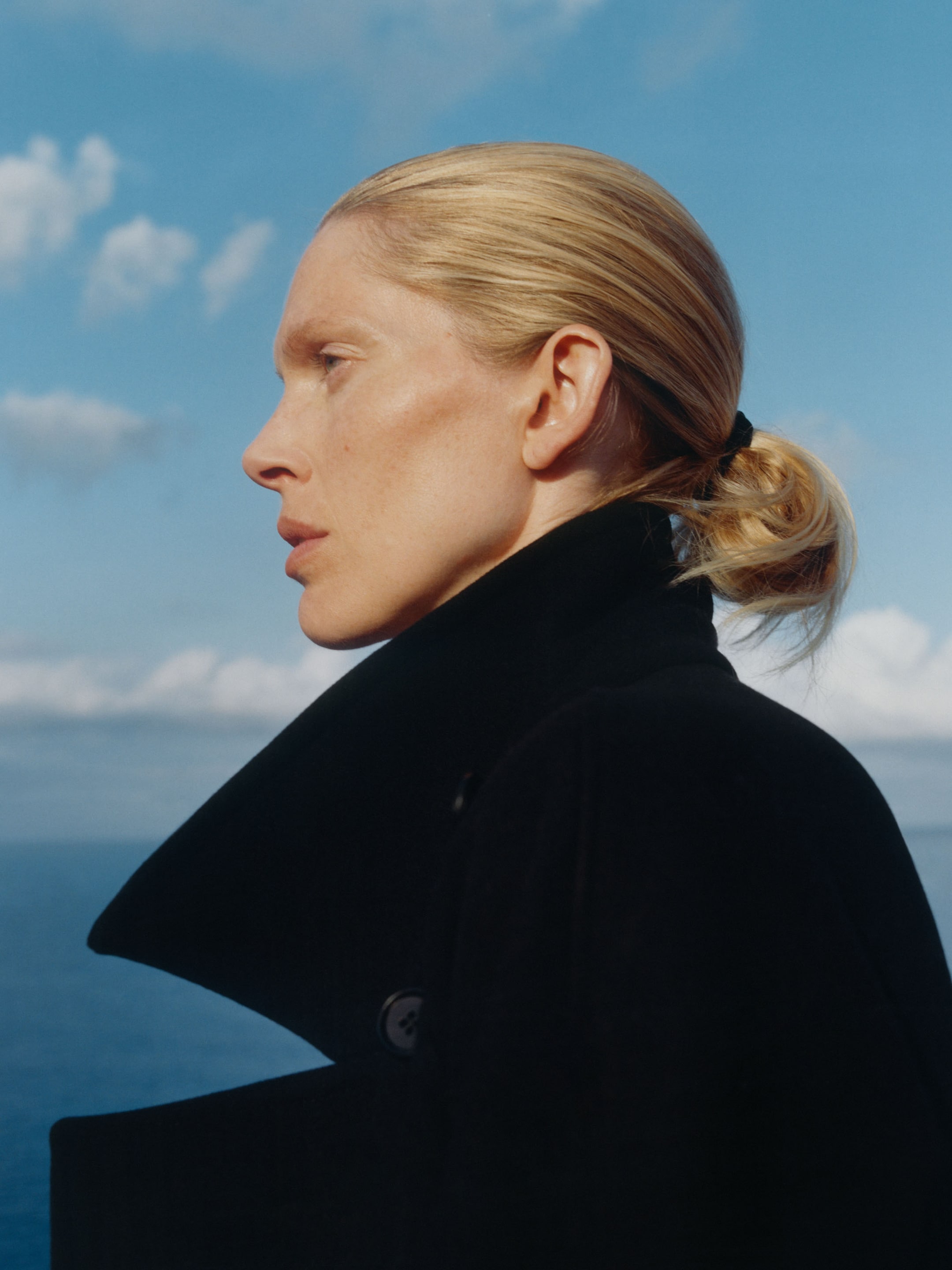 Side profile of a person with blonde hair tied back, wearing a dark coat, against a clear blue sky and ocean backdrop.