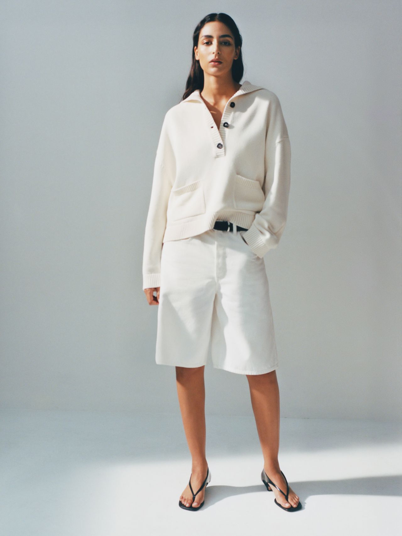 Woman in a white sweater and shorts stands against a light background, wearing black sandals, with hands in pockets, looking at the camera.