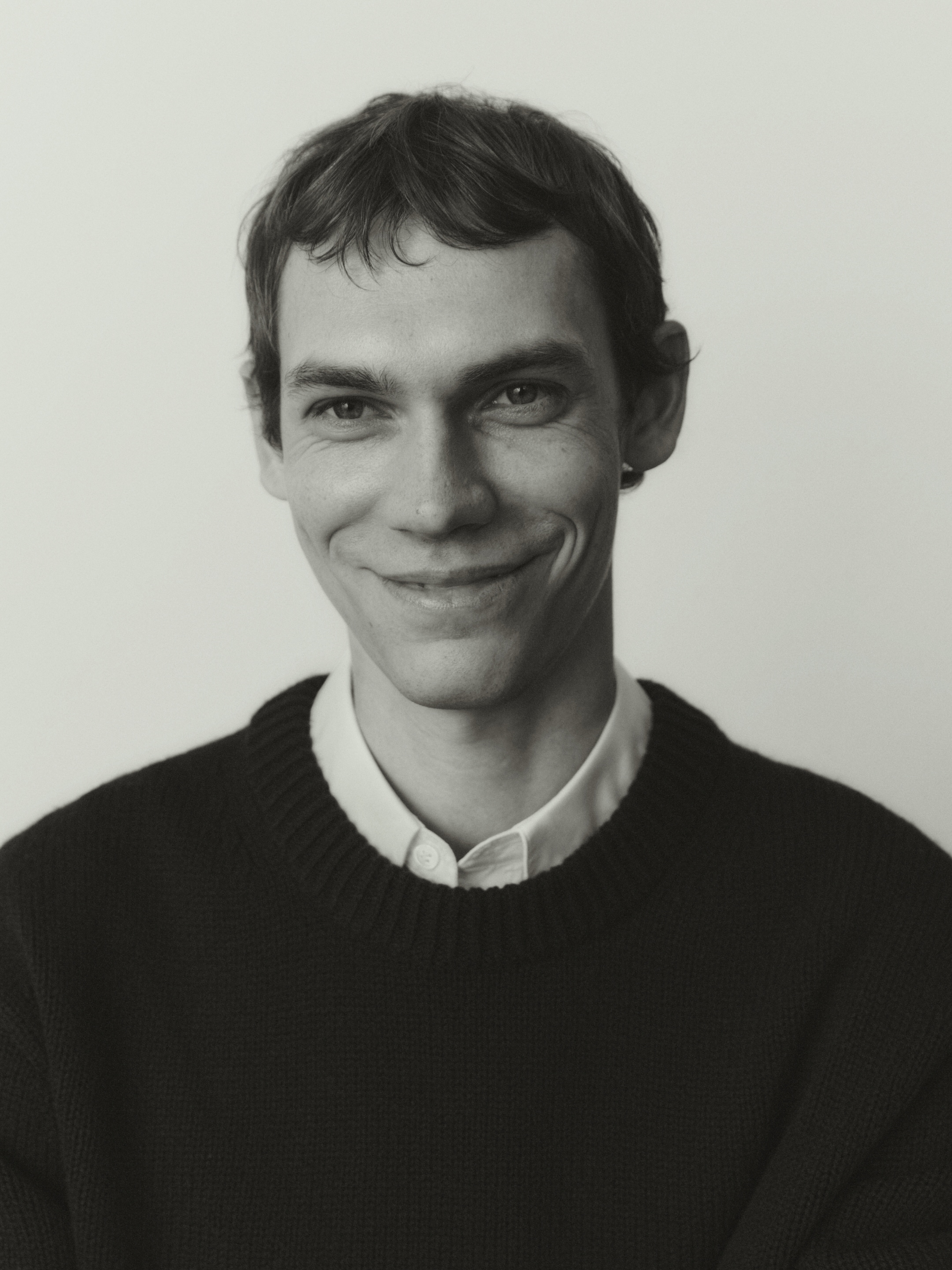 Black and white portrait of a smiling person wearing a dark sweater over a collared shirt.