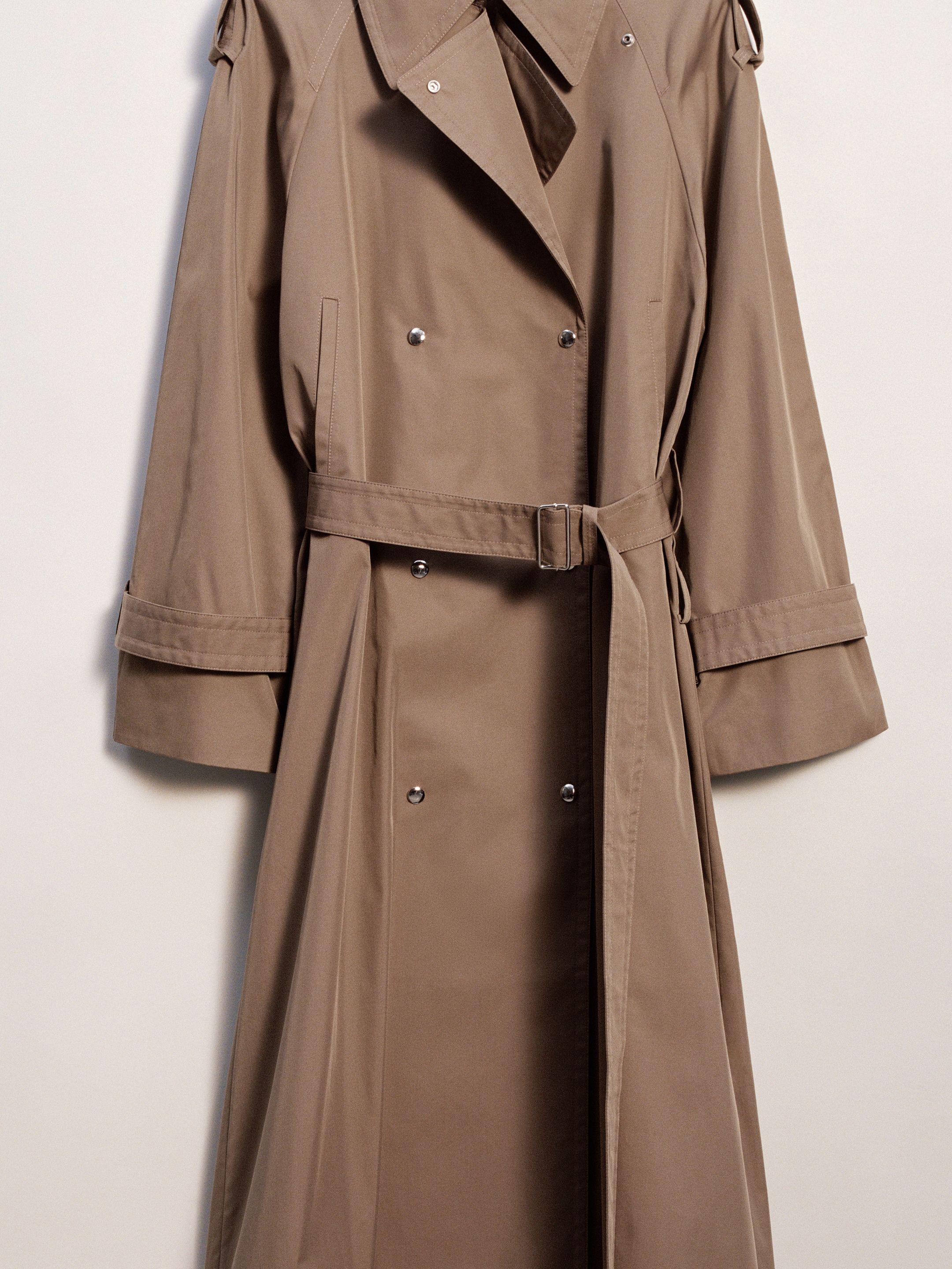 A beige trench coat with a belt, button details, and shoulder epaulettes against a plain background.