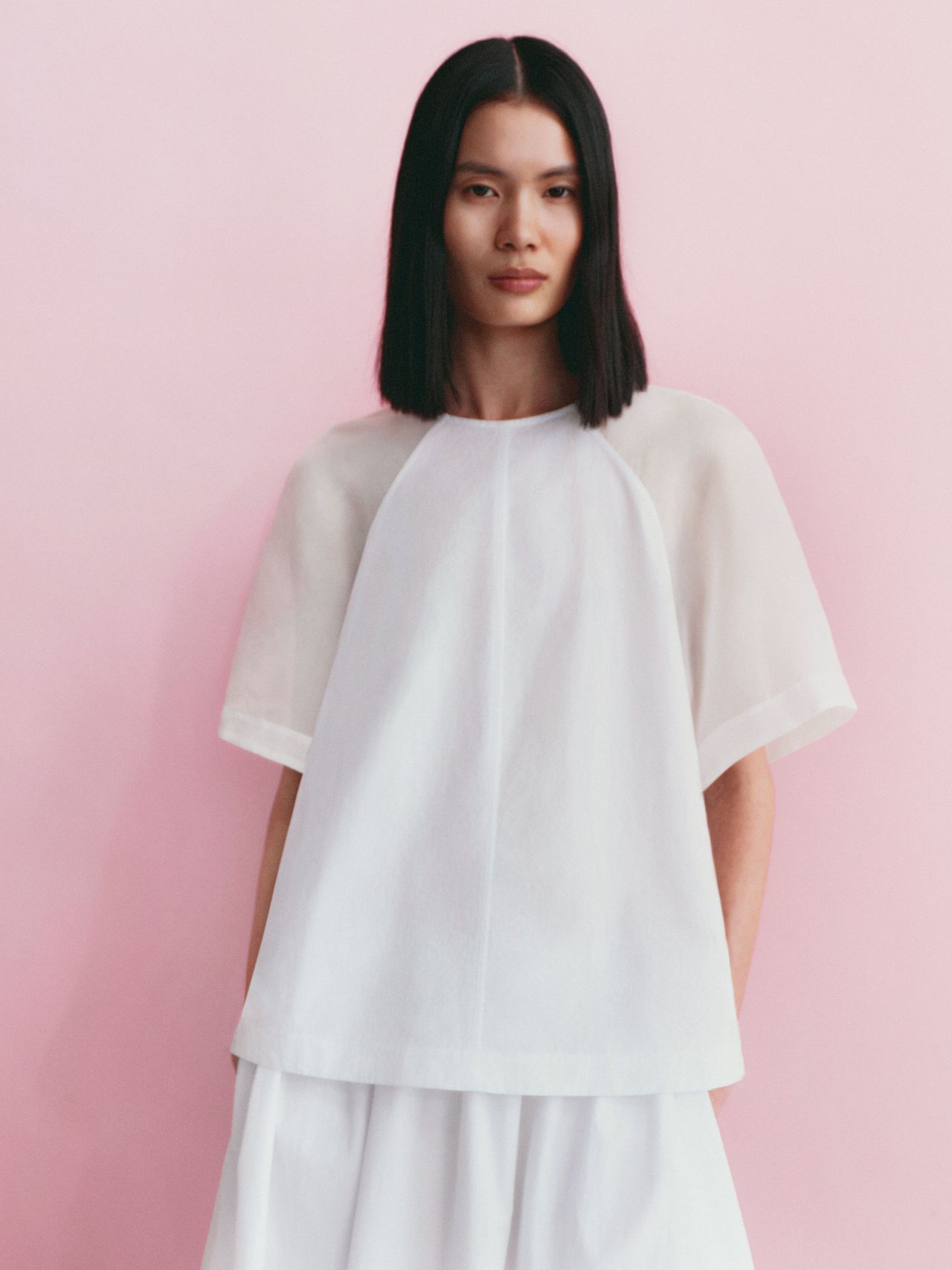 Woman in a white, sheer-sleeved top and skirt against a soft pink background.