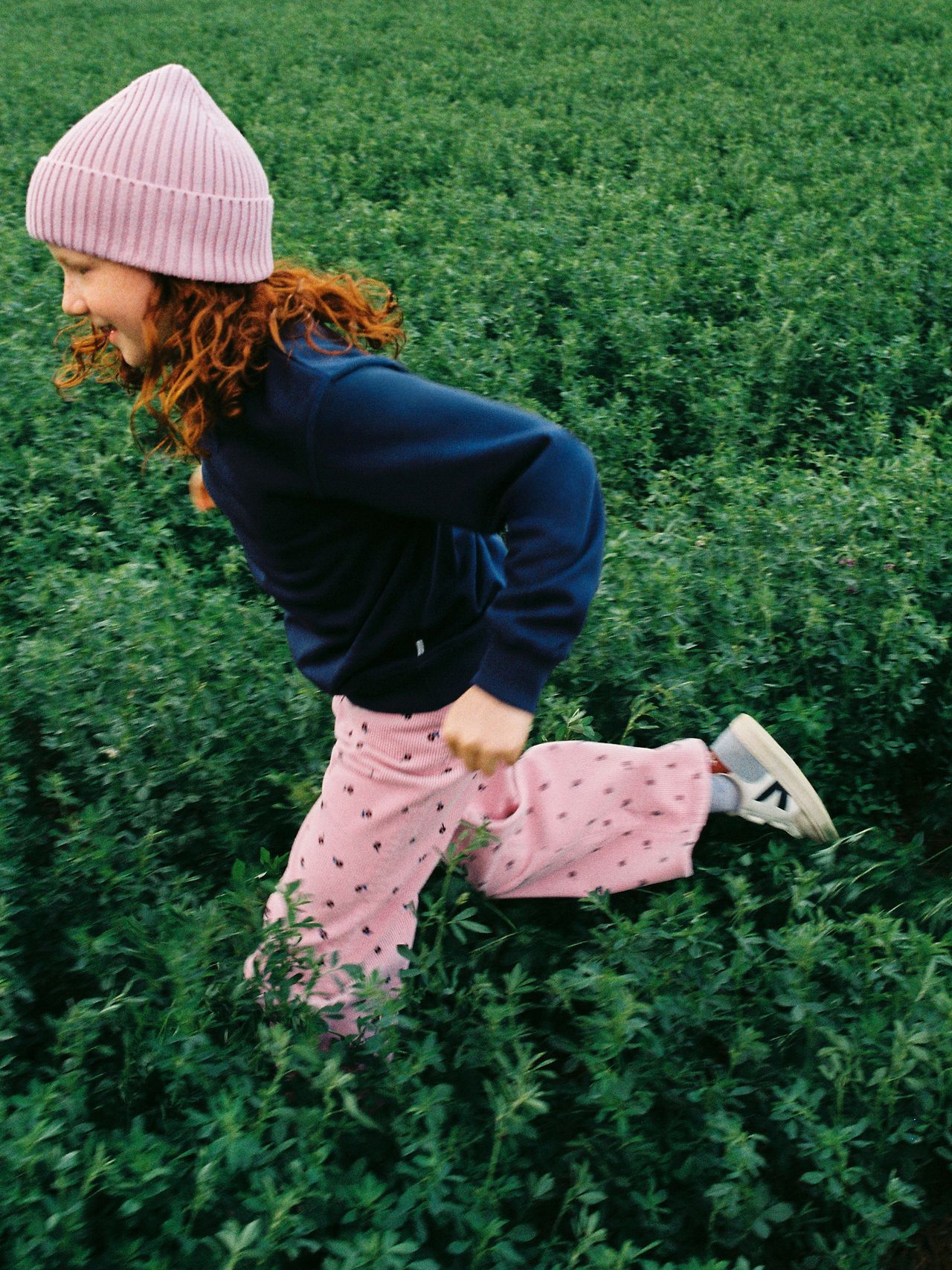 Child in a pink hat and pants runs joyfully through a lush green field, wearing a navy sweater and sneakers.