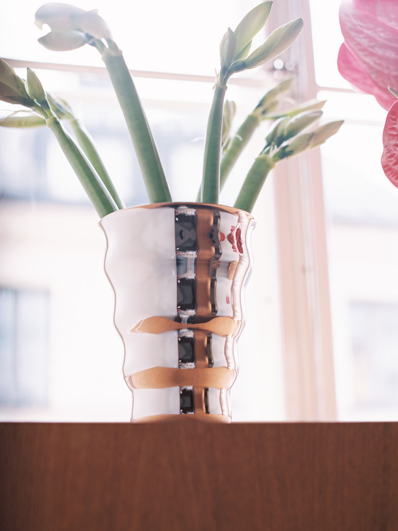 A modern, reflective vase with green stems on a wooden surface, placed in front of a window with blurred outdoor buildings visible.
