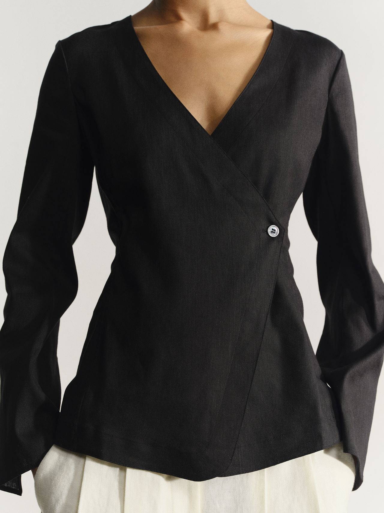 A person wearing a stylish black wrap top with long sleeves and a small button, paired with a white skirt, against a plain background.