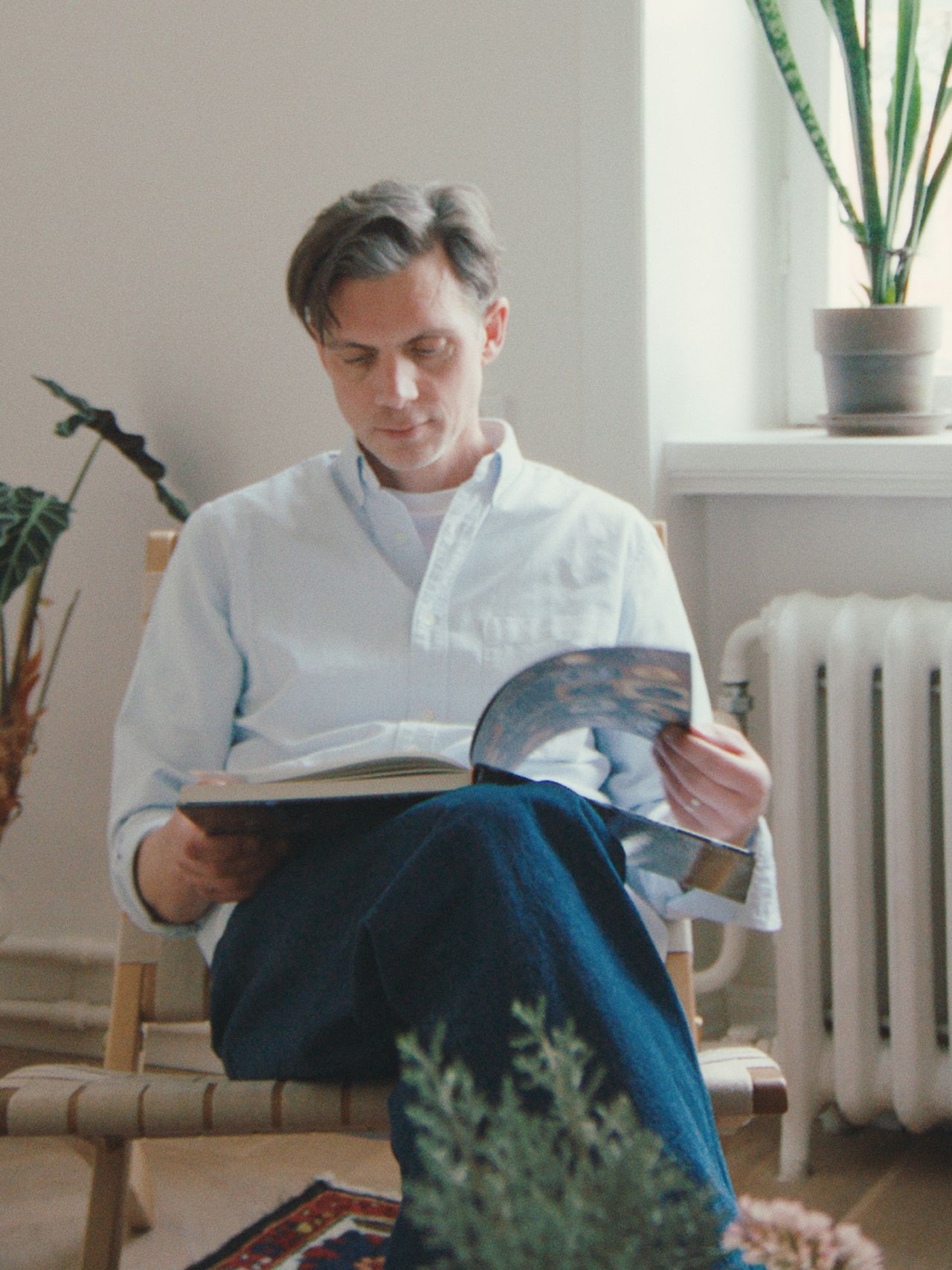 A person sitting indoors on a wooden chair, reading a book and a magazine, with potted plants nearby and natural light coming from a window.