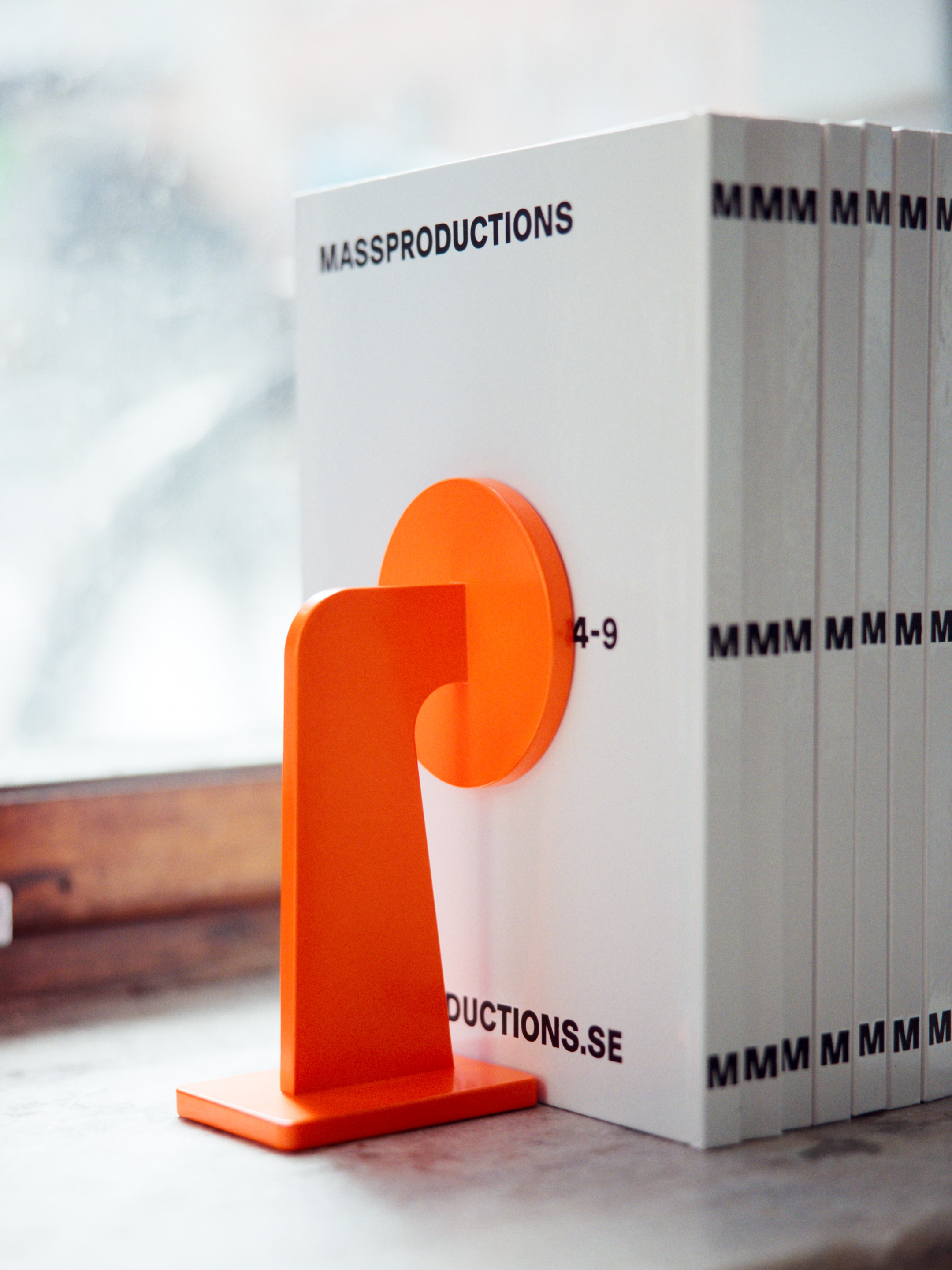 Orange bookend holding up a row of white books with "MASSPRODUCTIONS" and "MMM" on the spines, placed on a windowsill.