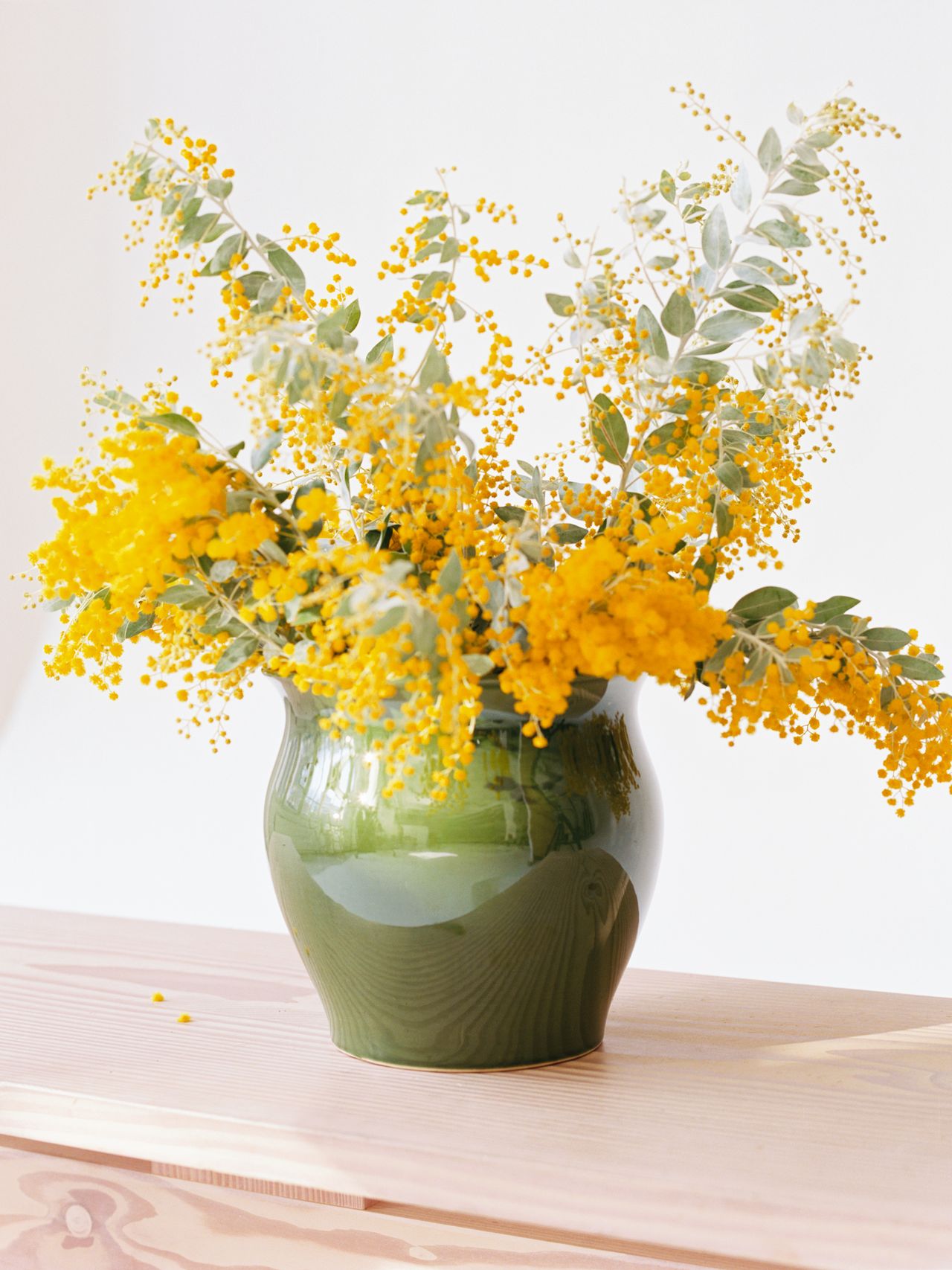Green vase with vibrant yellow flowers and silvery-green leaves on a light wooden surface, against a soft white background.