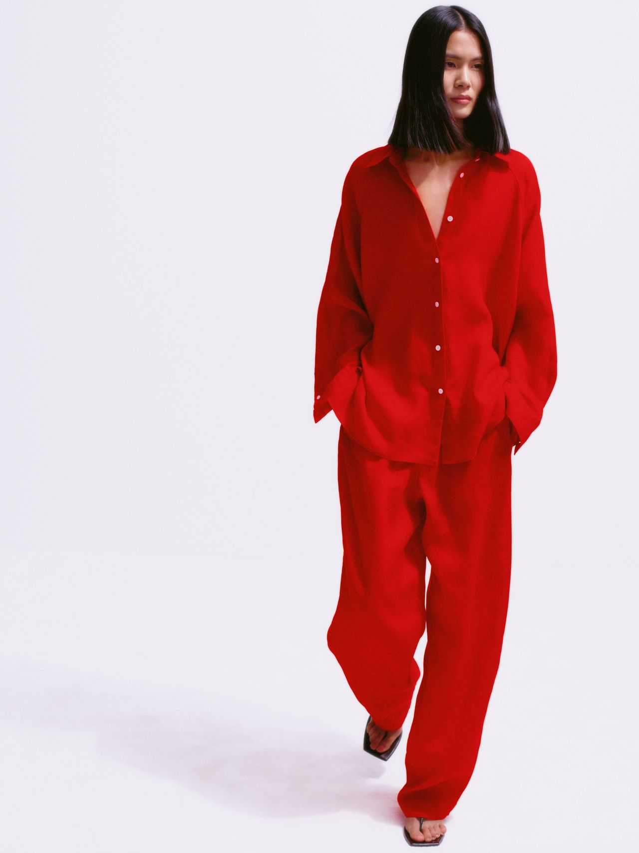 A woman with long hair wears a red, loose-fitting shirt and pants, and black sandals, walking against a plain white background.