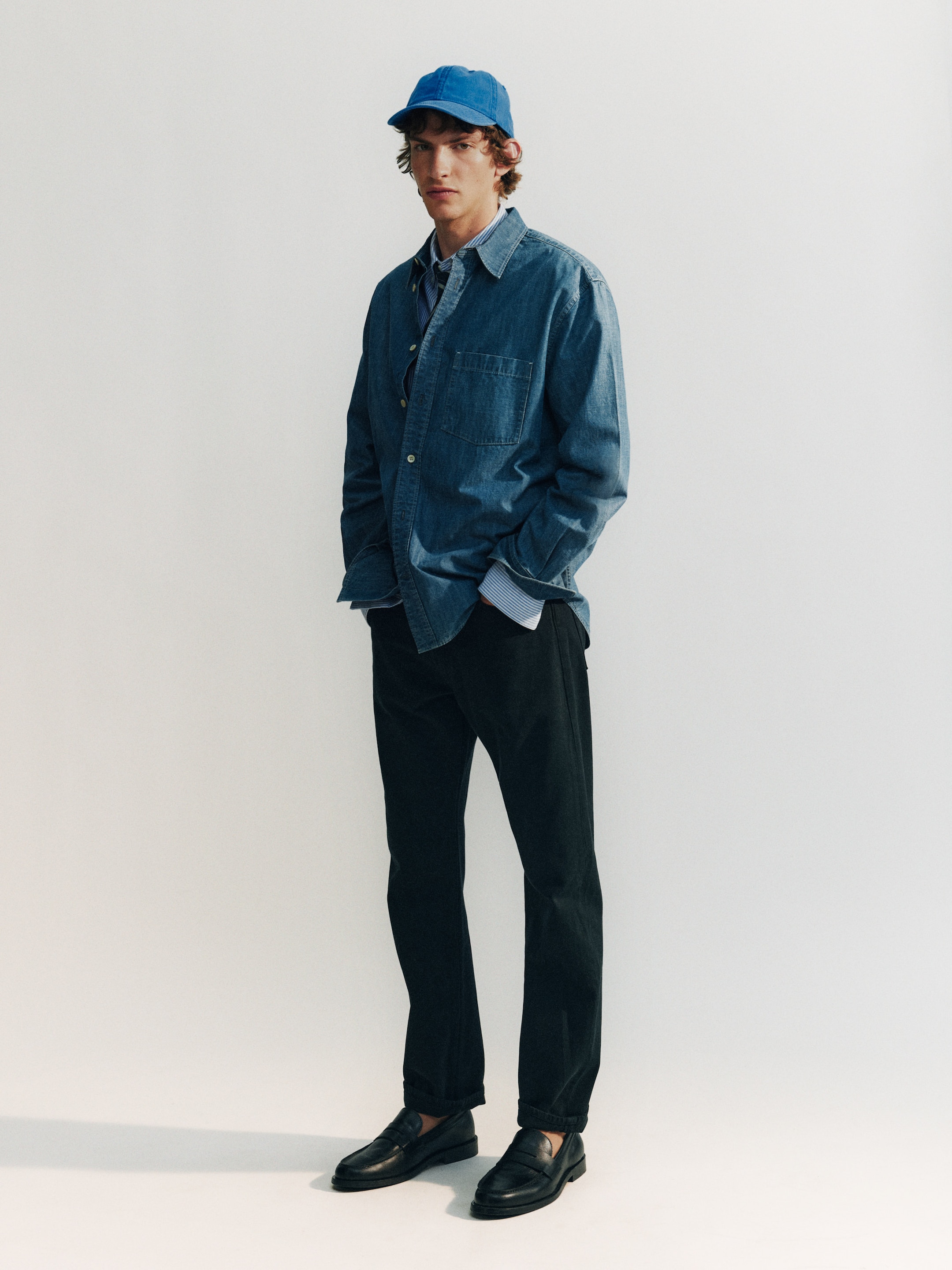 Man in a blue denim shirt and cap, wearing dark pants and black loafers, stands against a plain white background with hands in pockets.