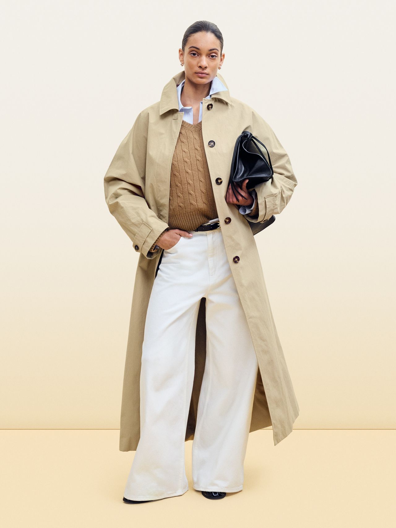 Person wearing a beige trench coat, brown sweater, white wide-leg pants, and holding a black clutch against a light background.