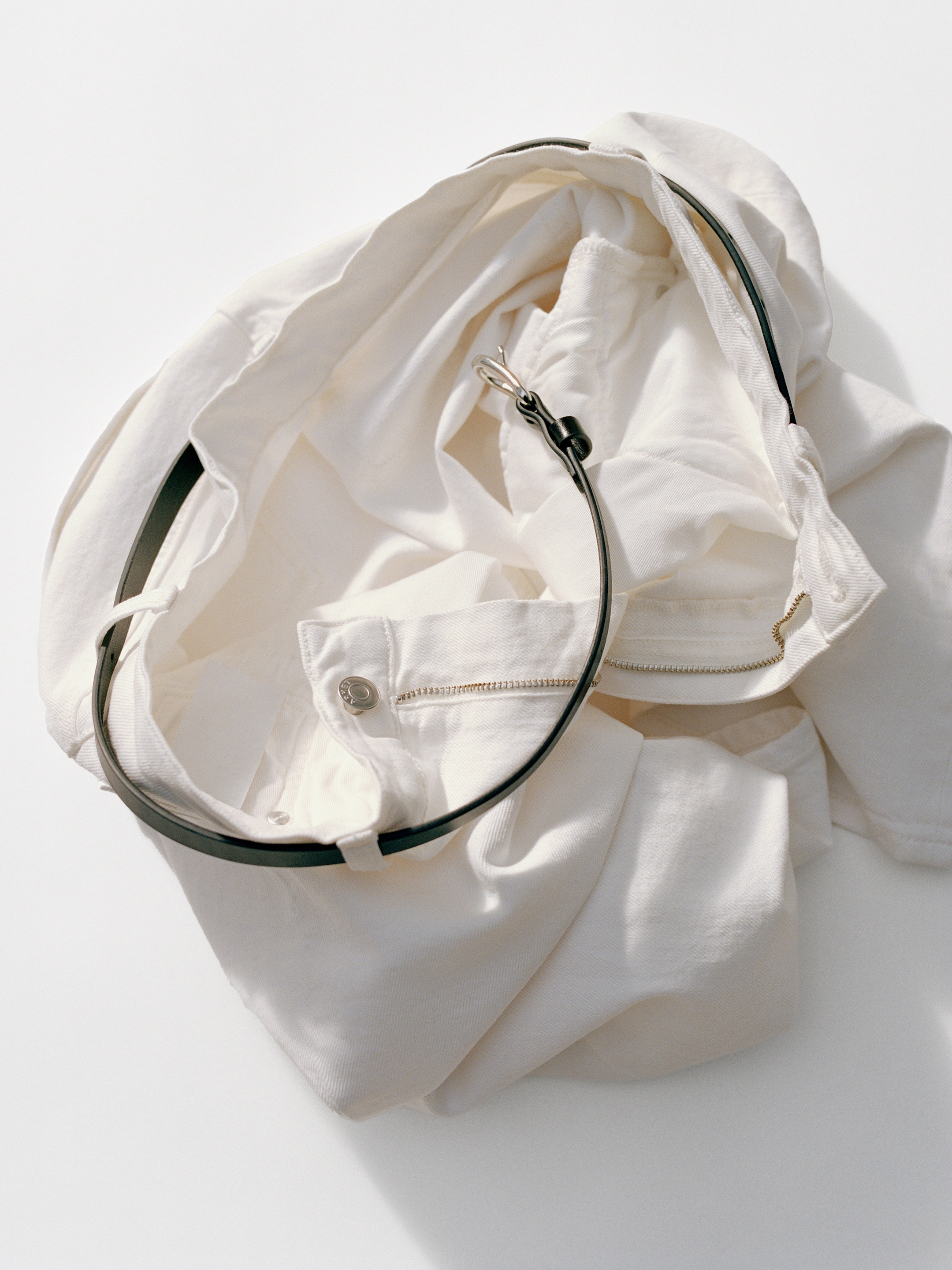 White pants crumpled on a surface, featuring a silver belt looped through the waistband and a visible silver button.