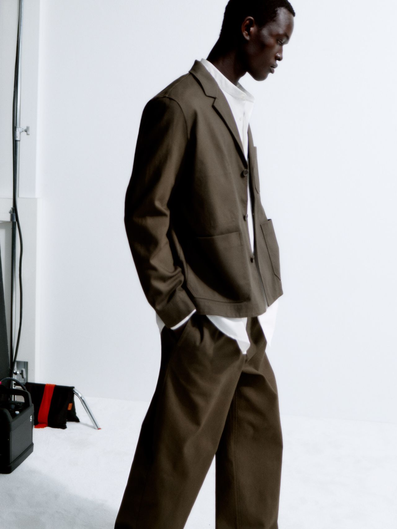 Man in a brown suit and white shirt stands against a white background, hands in pockets, with studio equipment in the corner.