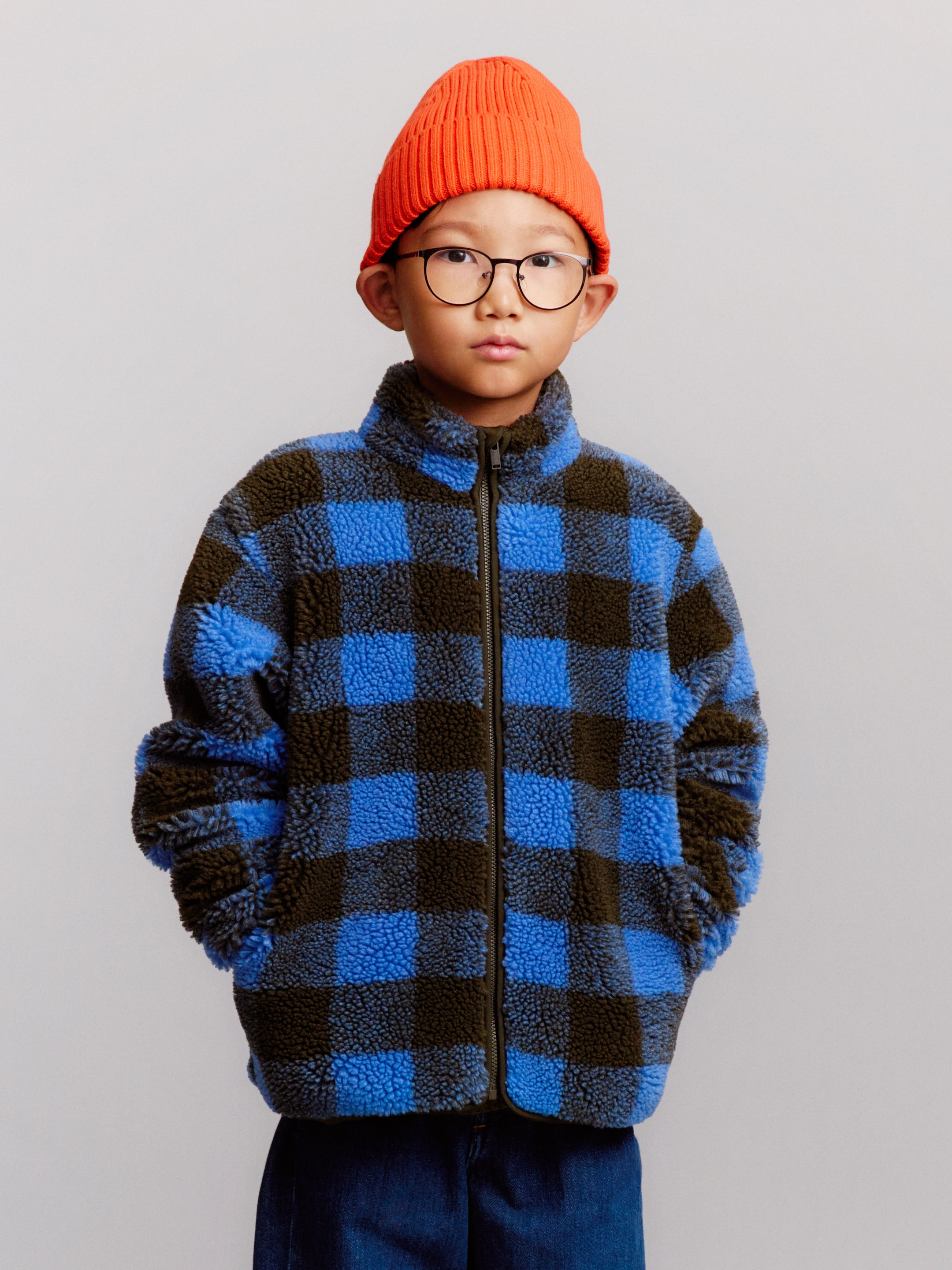 A child wearing glasses, an orange beanie, and a blue and brown checkered fleece jacket stands against a neutral background, hands in pockets.