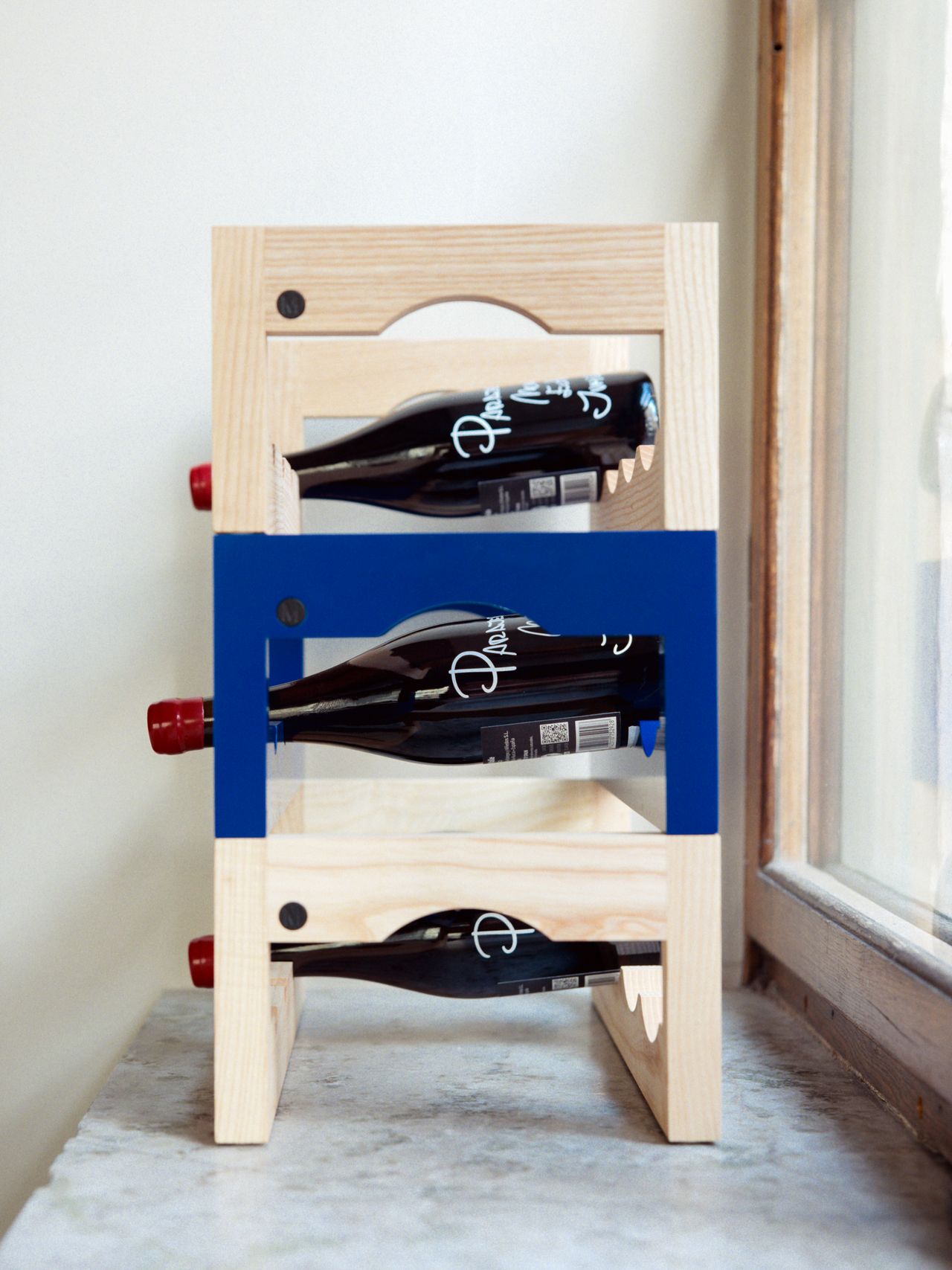 Wooden wine rack holding three bottles with red caps, positioned next to a window on a stone surface.