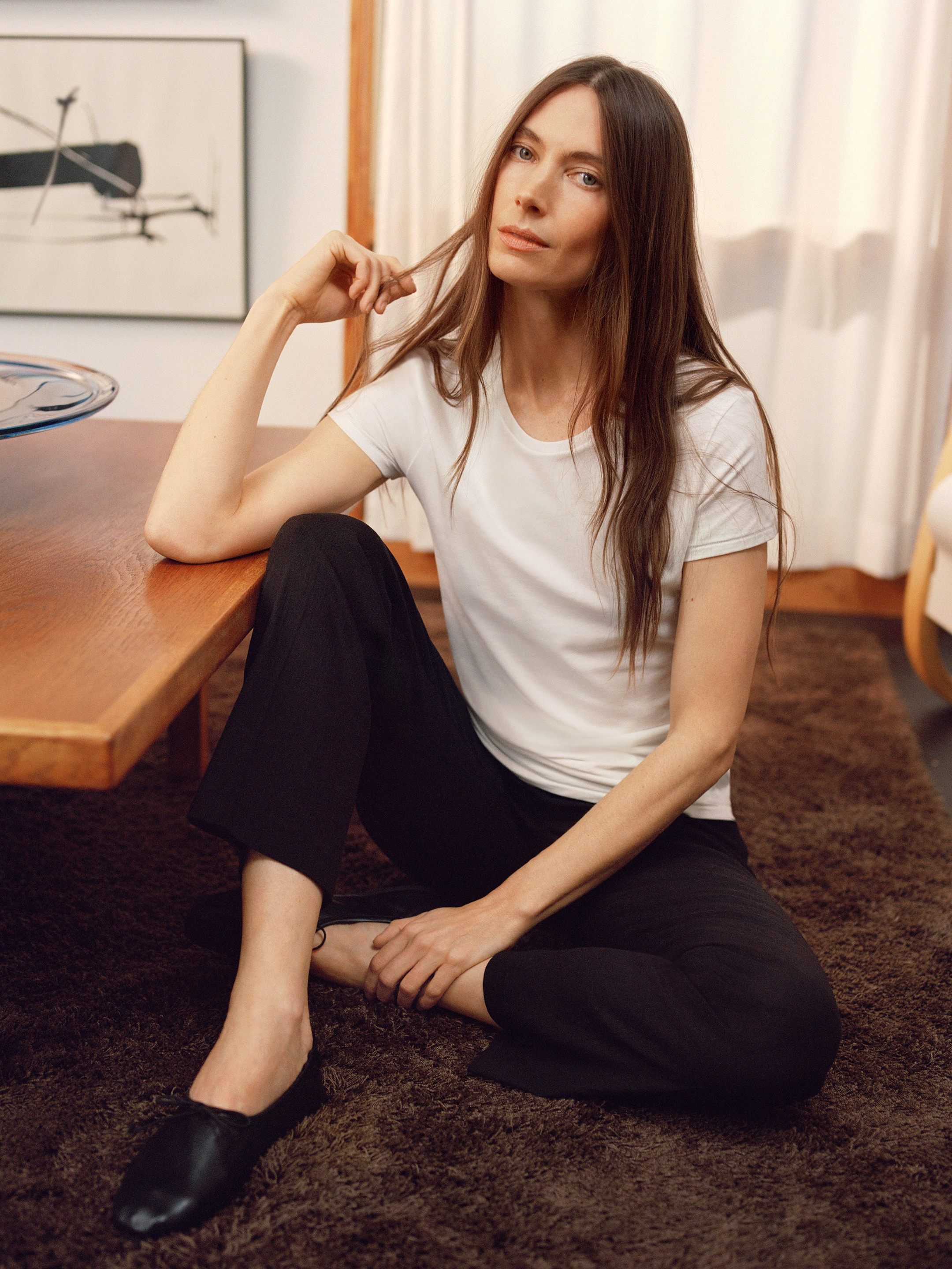 A person with long hair sits on a brown carpet, wearing a white shirt and black pants, resting their elbow on a wooden table.