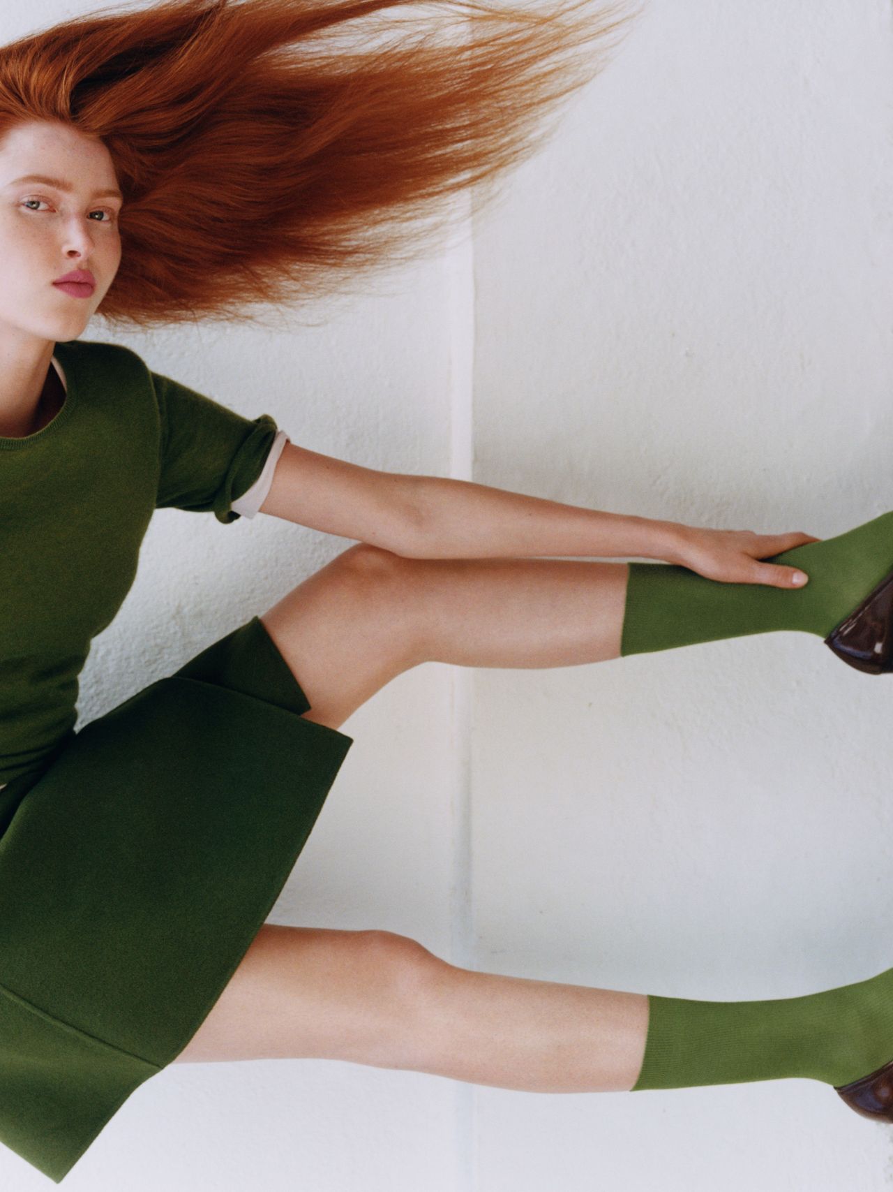 Person in a green outfit poses against a white background, with long red hair flowing outward, creating a dynamic and stylish composition.