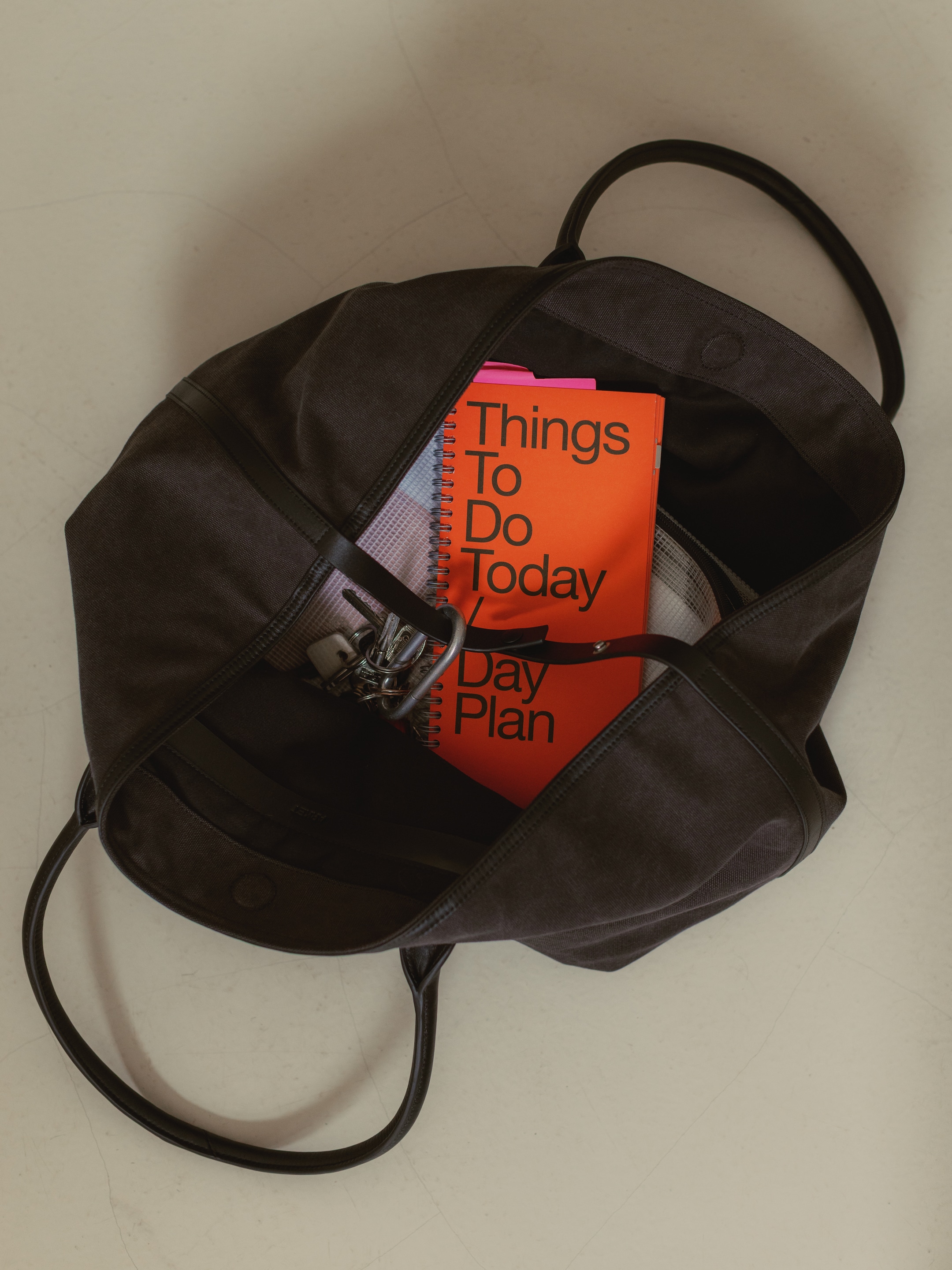 Open black bag containing a bright orange notebook titled "Things To Do Today," with keys and a rolled-up magazine inside.