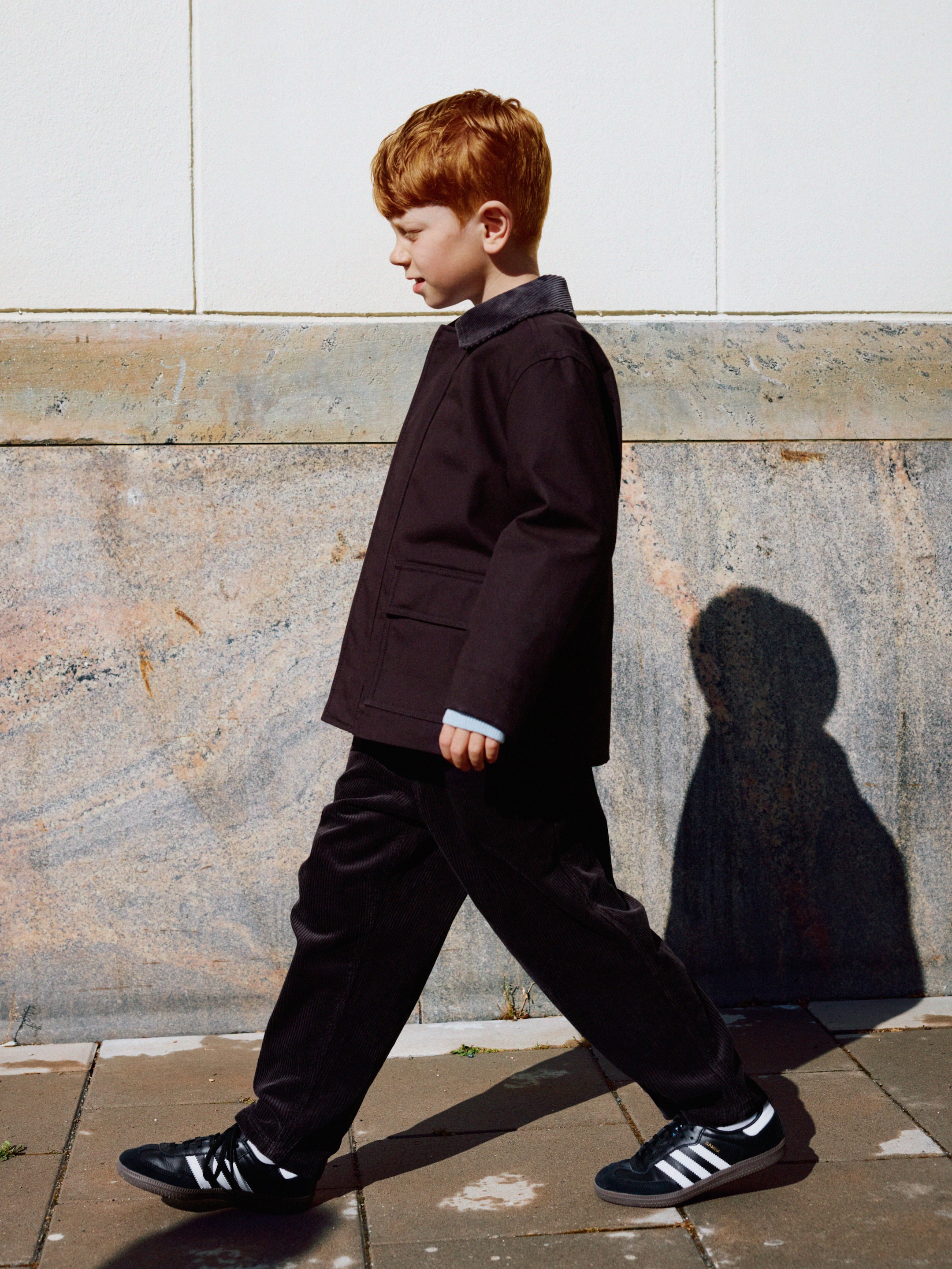 A young boy with red hair walks on a tiled path, wearing a dark coat and sneakers, casting a shadow against a stone and white wall.