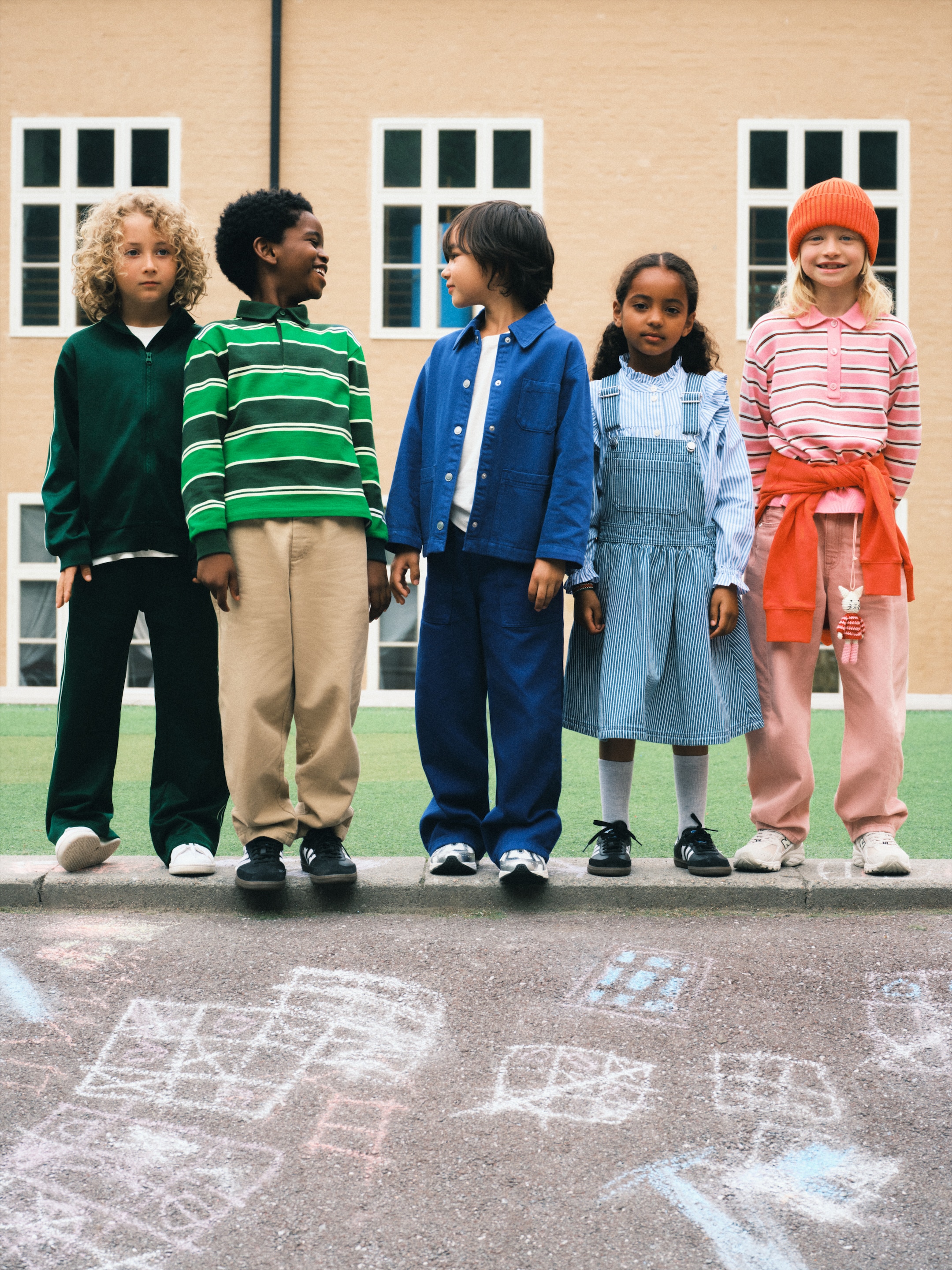 Five children stand in front of a building on a playground with chalk drawings on the ground, dressed in colorful outfits.