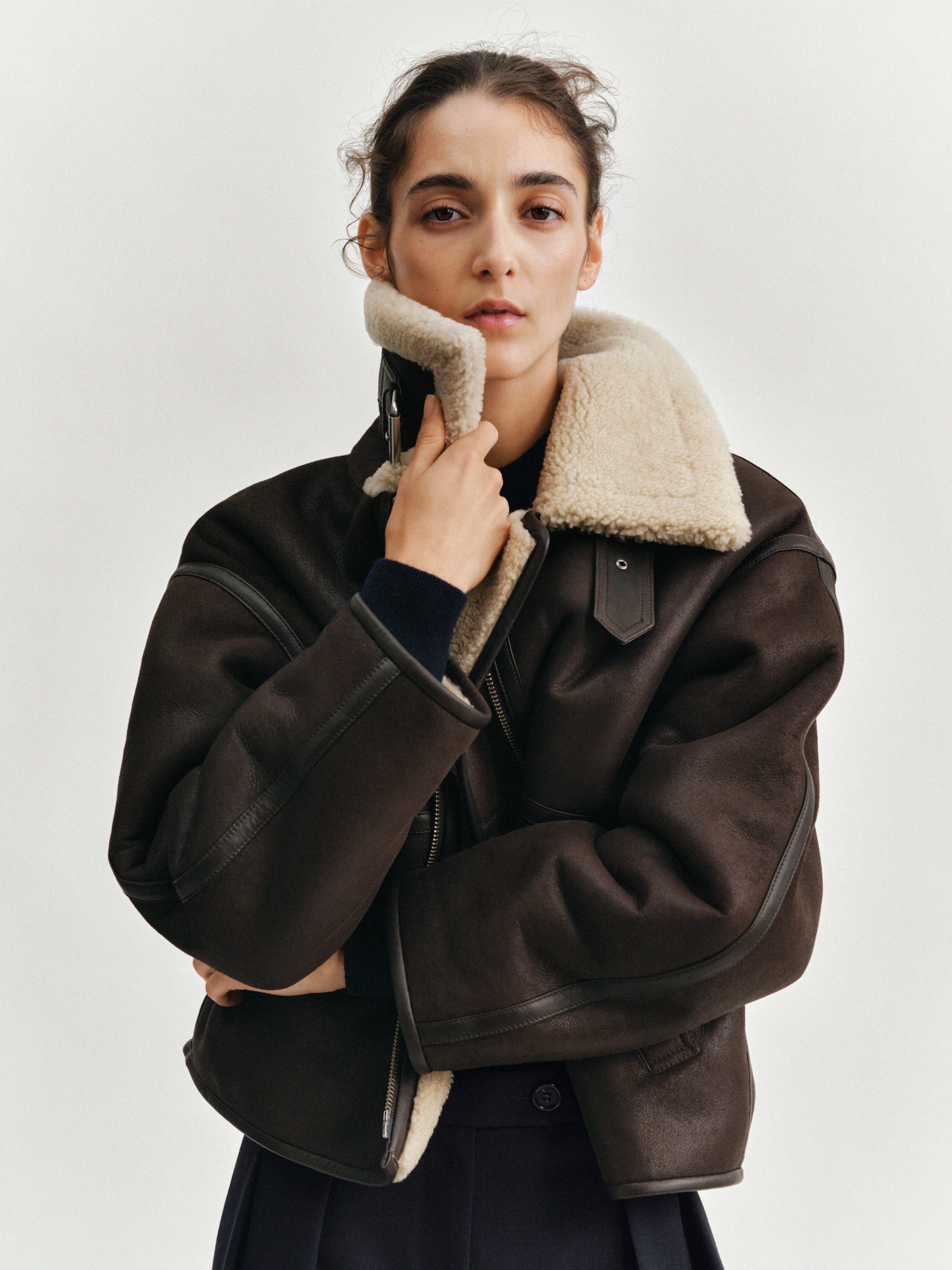 A person wearing a large dark brown shearling jacket with a light beige fur collar, looking directly at the camera.